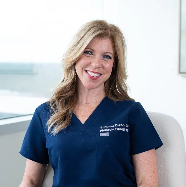 Woman in blue medical scrubs sitting in an office, smiling at the camera.