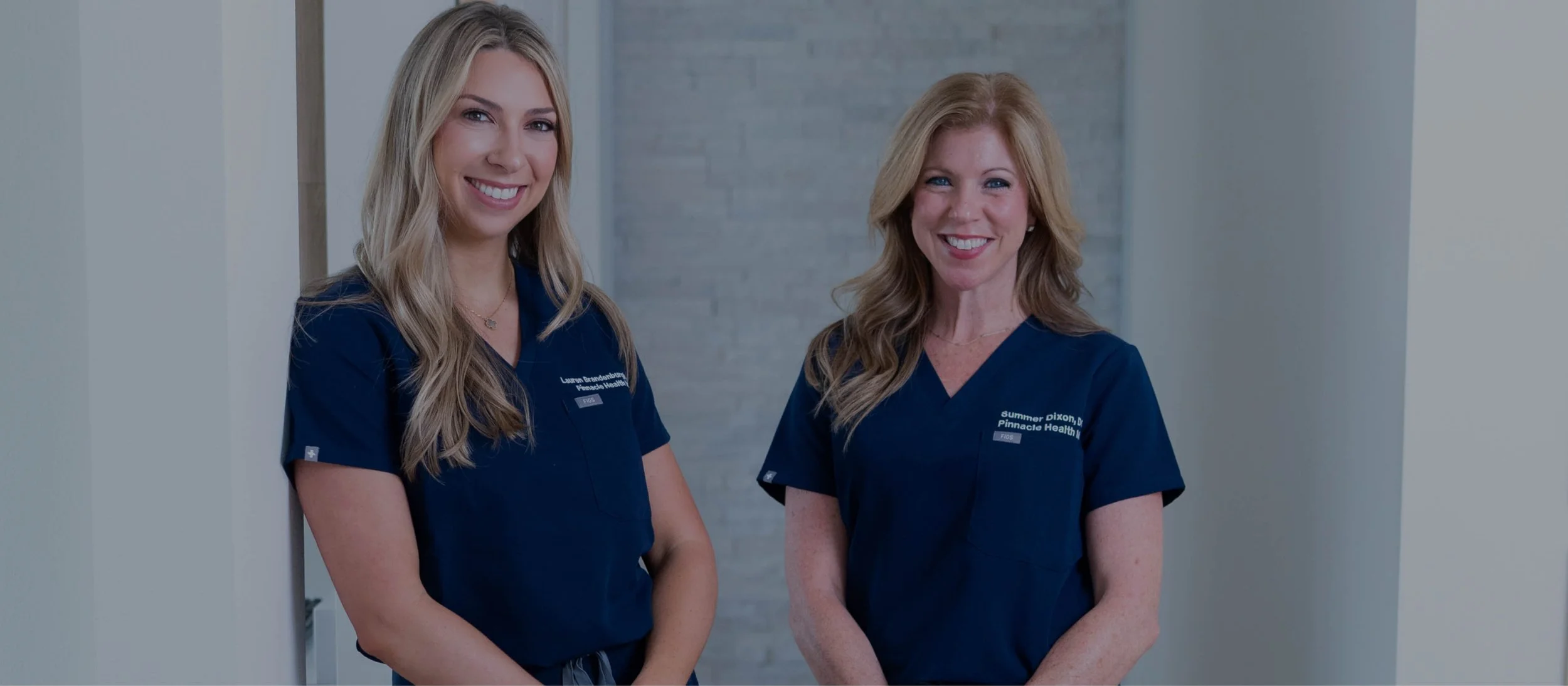 Two women wearing navy blue scrubs standing against a light-colored wall, smiling at the camera.