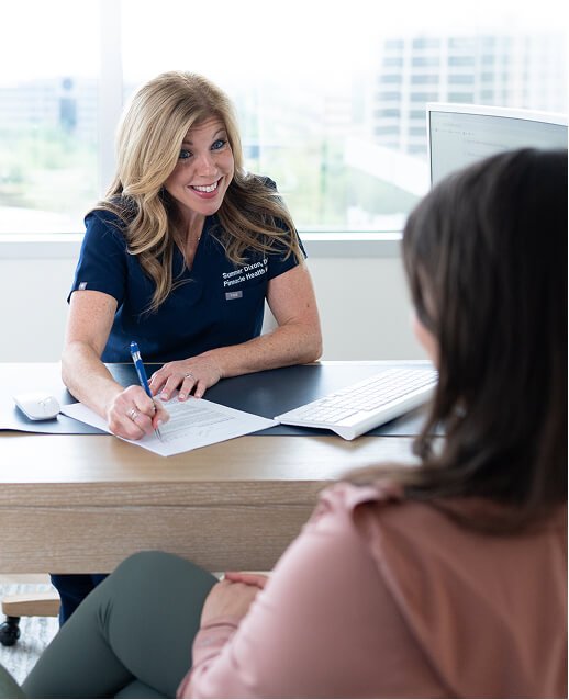 A woman in a navy blue shirt sitting at a desk, talking to another woman with brown hair, in an office with large windows and a cityscape background.