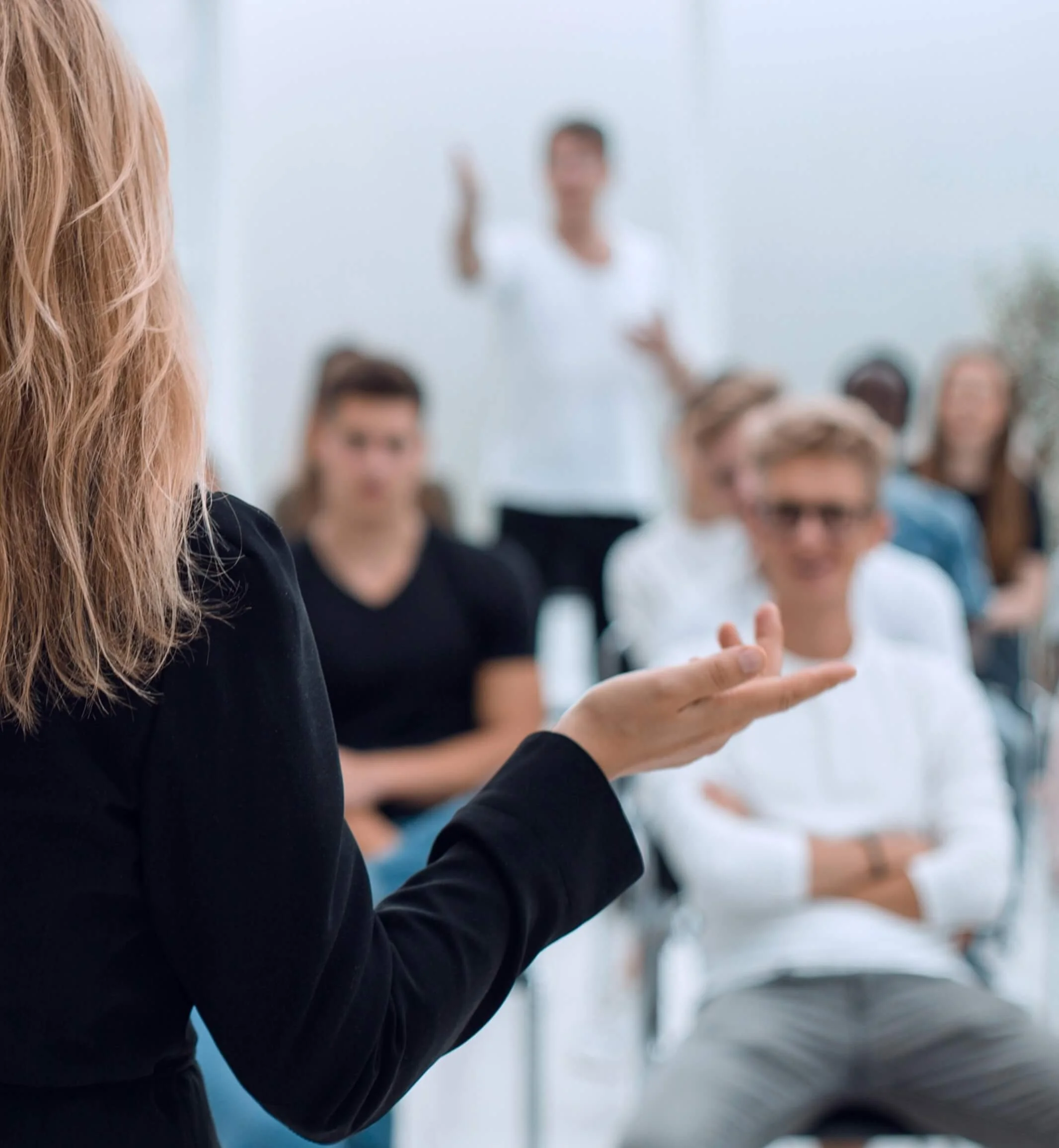 A woman with long red hair wearing a black blazer appears to be speaking or presenting in front of an audience, with several blurred individuals in the background.