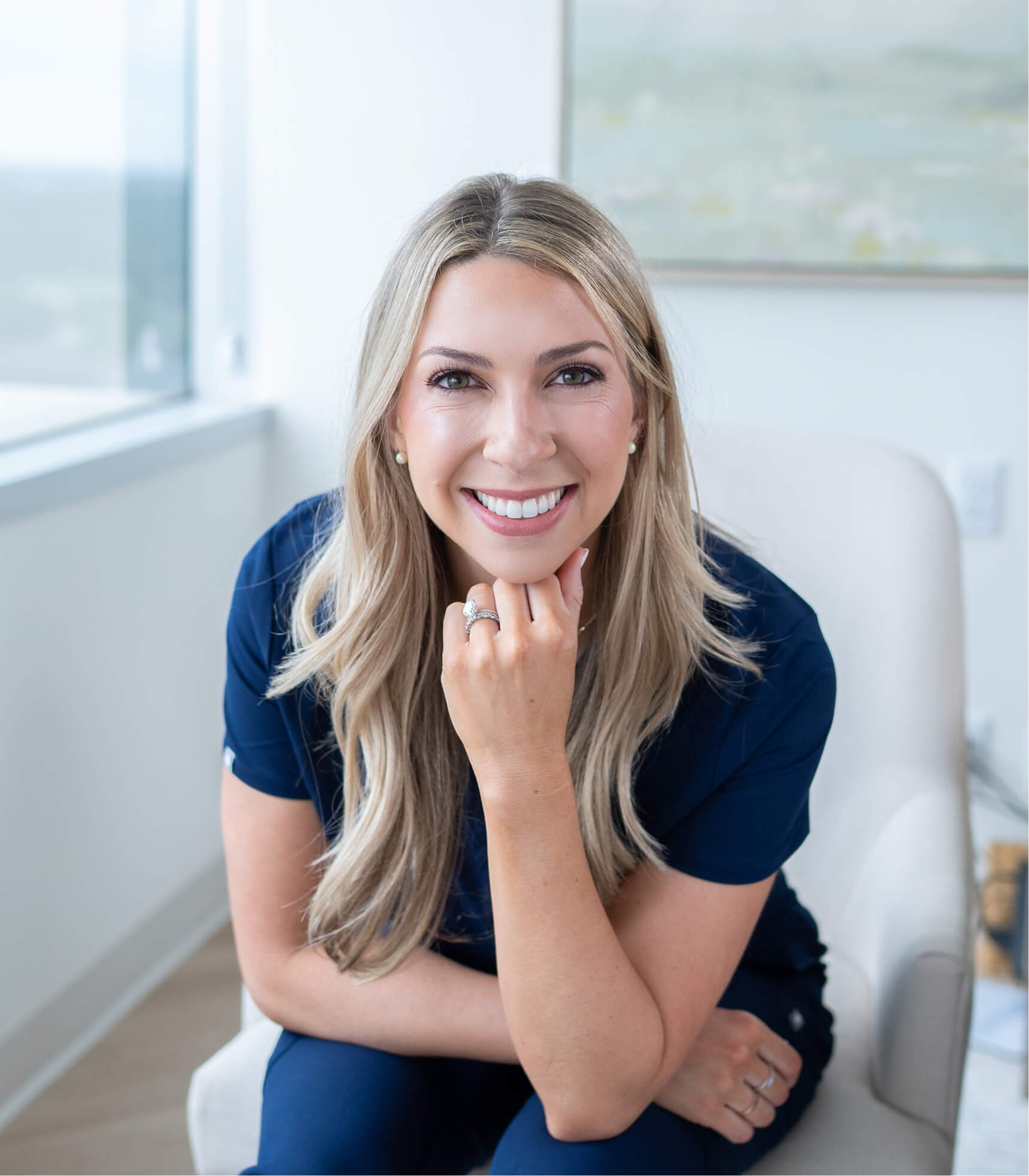 A woman smiling, sitting on a white chair near a window with a view of trees, wearing a blue outfit, with long blonde hair and jewelry.