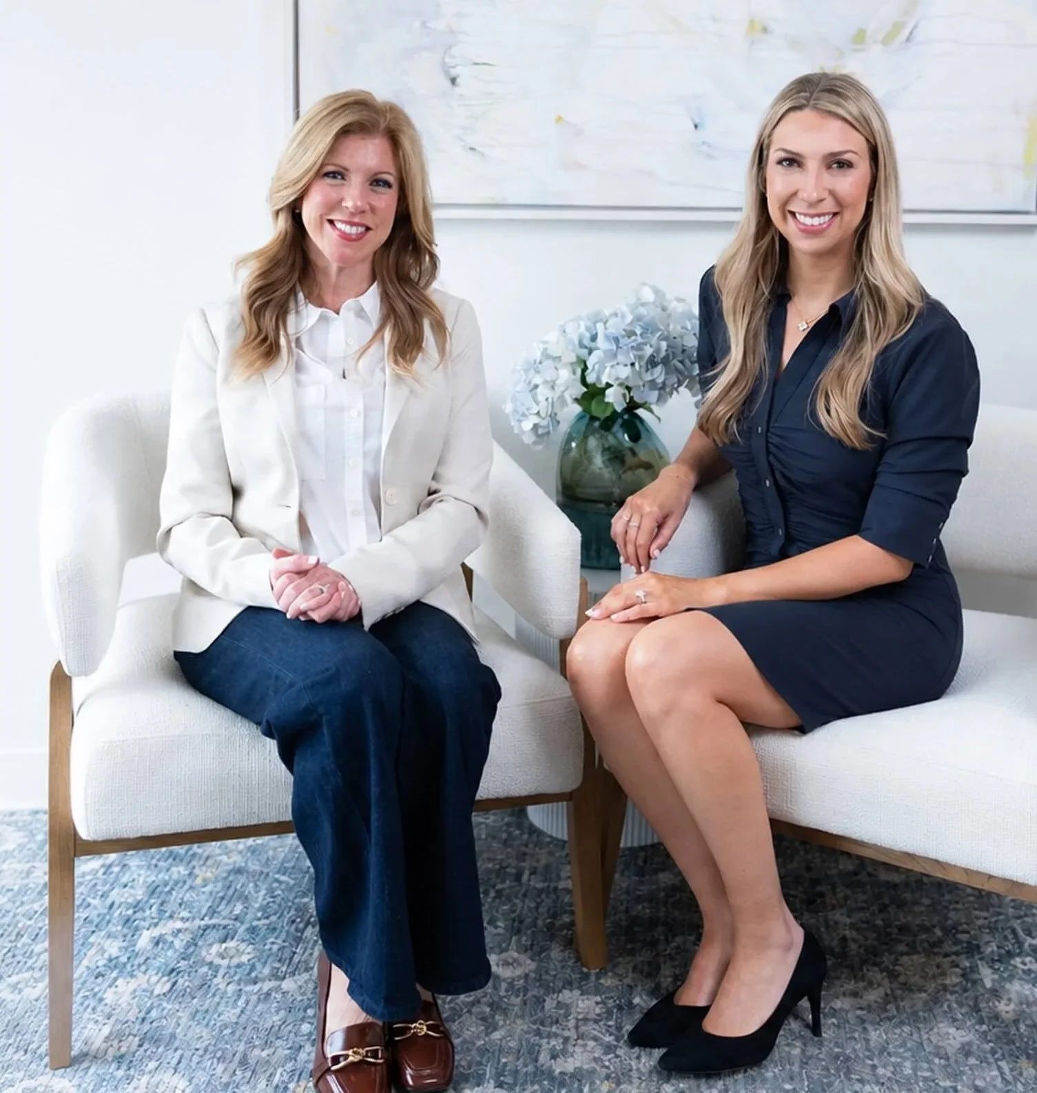 Two women sitting on a white sofa, smiling, with a glass vase of white flowers on a side table behind them. One woman wears a white blazer and dark pants, the other a navy blue dress and black heels. A large abstract painting hangs on the wall behind them.