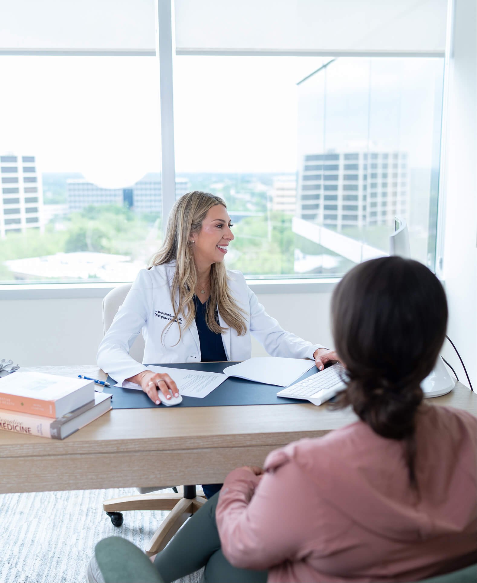 A female doctor with long blonde hair, wearing a white coat, smiling and talking to a patient in an office with large windows overlooking a cityscape with tall buildings.