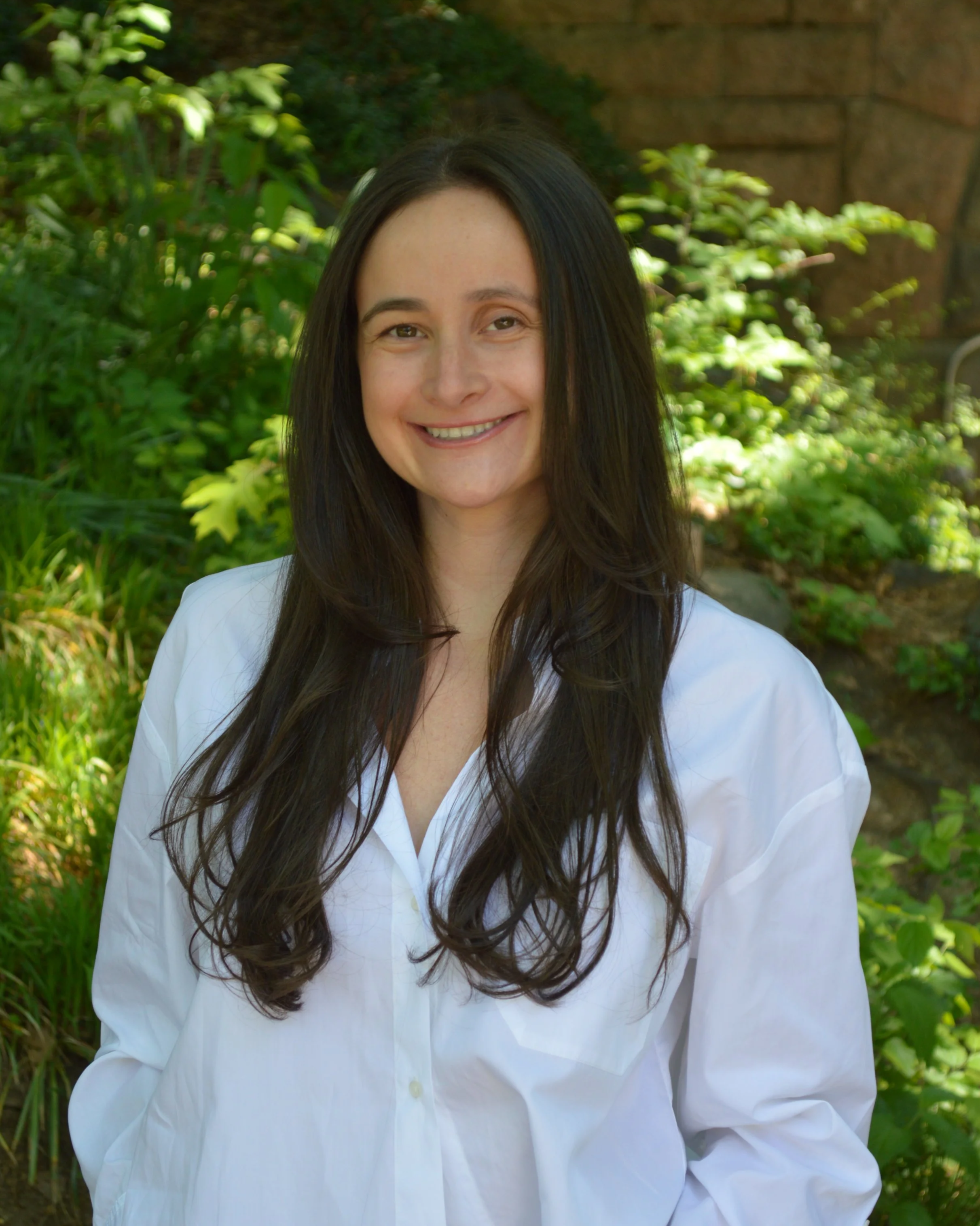 Smiling woman with long dark hair wearing a white shirt, standing outdoors with green plants and trees in the background, and a stone structure visible behind her.