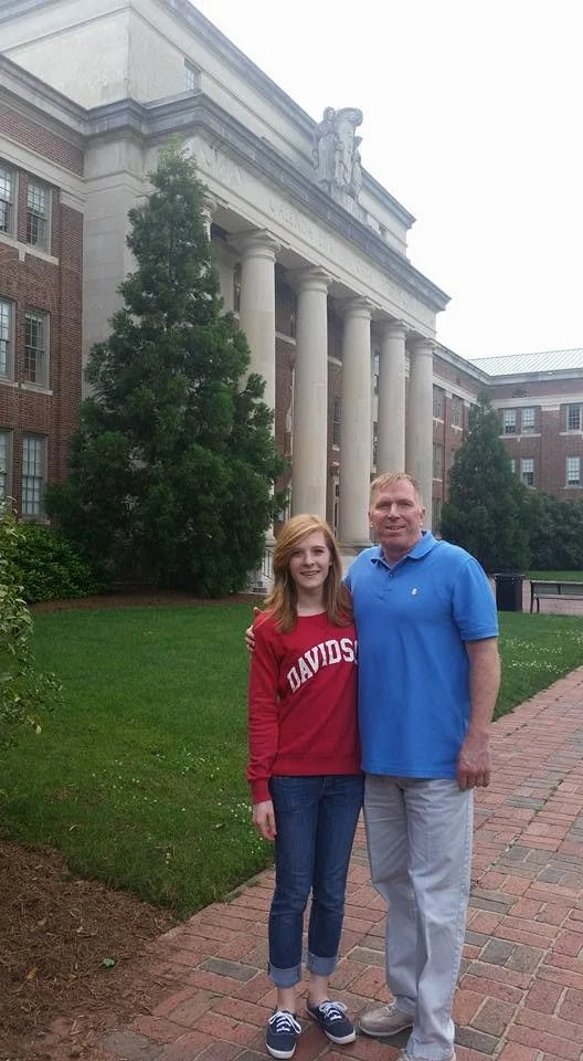 A young woman and an older man standing on a brick path in front of a large building with columns and a statue on the roof. The woman is wearing a red Davidson sweatshirt and jeans, and the man is wearing a blue polo shirt and light-colored pants.