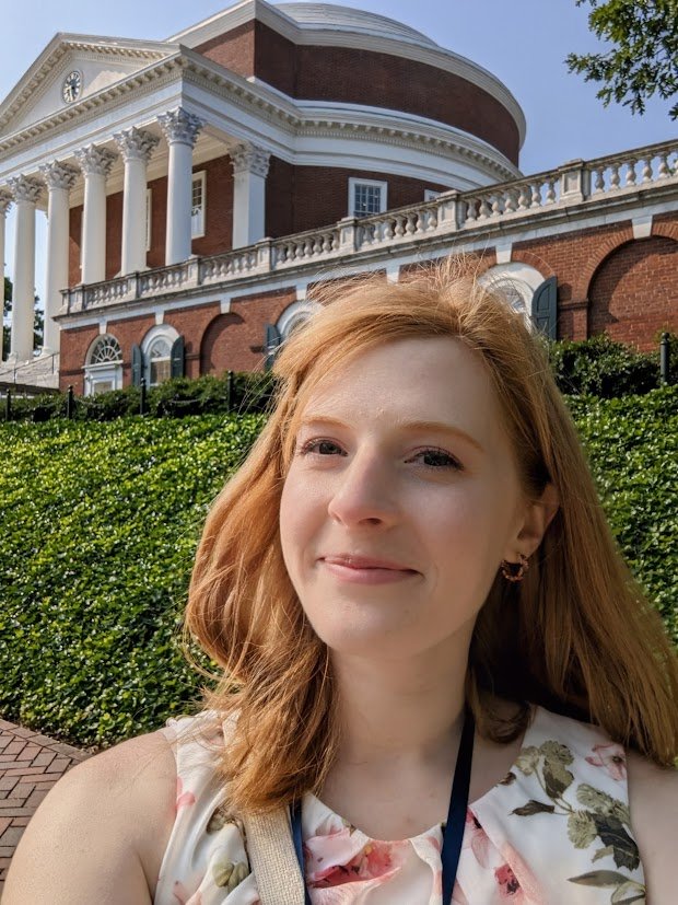 A young woman with red hair taking a selfie outdoors in front of a large, historic brick building with white columns and detailed architecture.