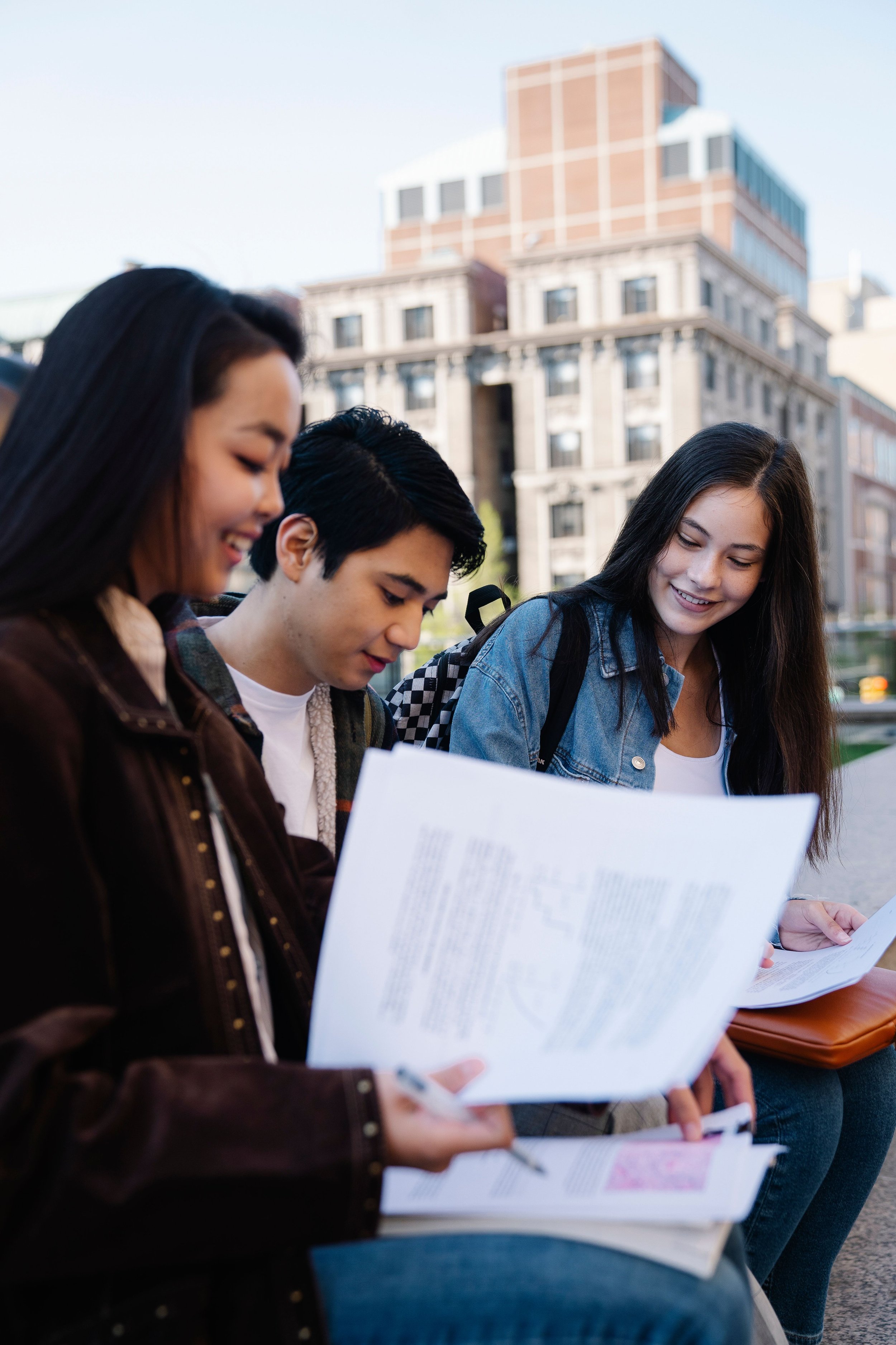 Four young students sitting outdoors, looking at papers and smiling, with buildings in the background.