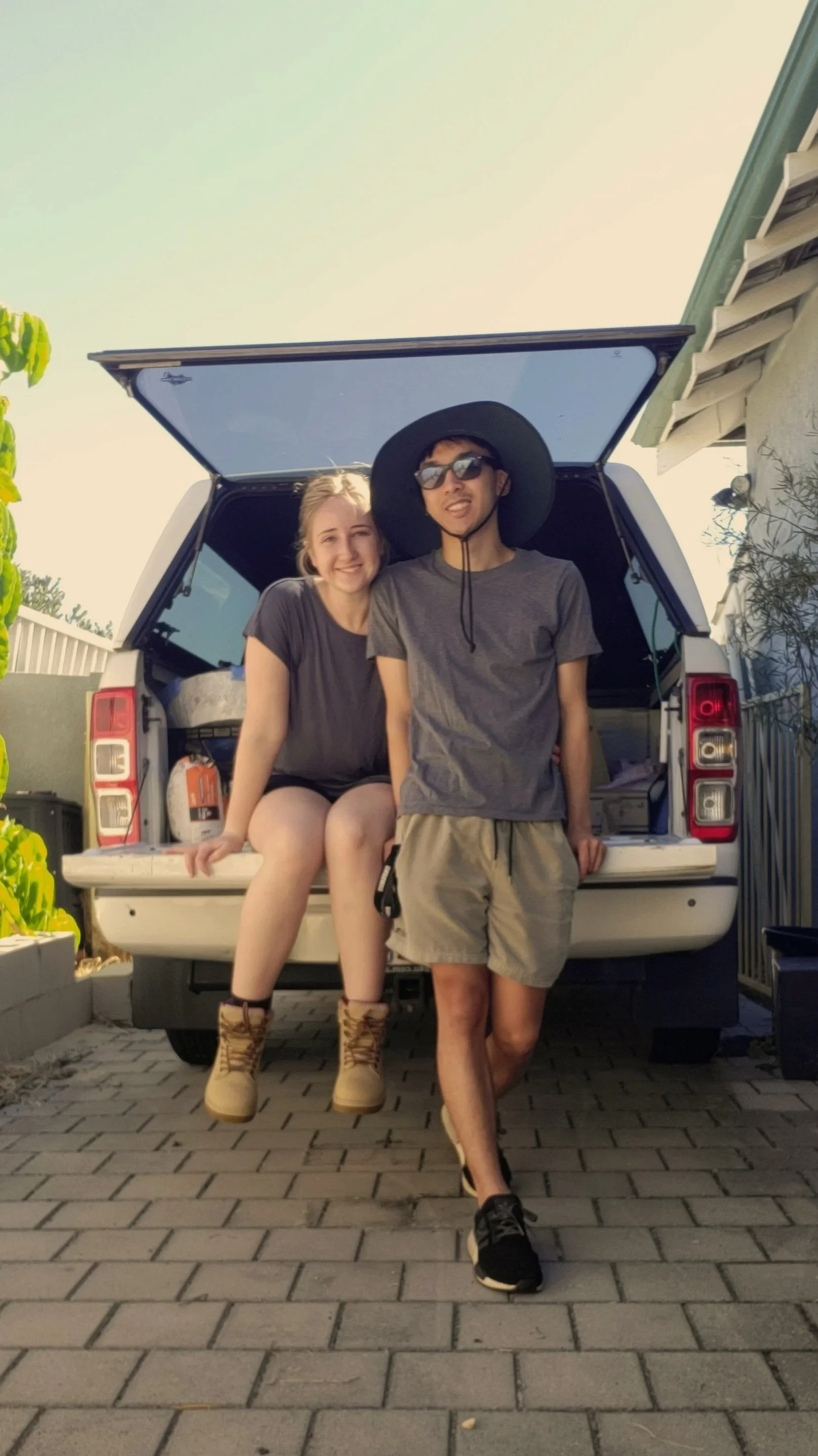 Two young people, a girl and a boy, are posing in the back of a white SUV with the tailgate open. The girl is sitting on the edge of the tailgate, smiling, wearing a dark T-shirt, shorts, and tan boots. The boy is standing on the ground, leaning against the SUV, wearing sunglasses, a wide-brimmed hat, a gray T-shirt, shorts, and sneakers. They are outdoors on a paved surface near a house and some greenery.