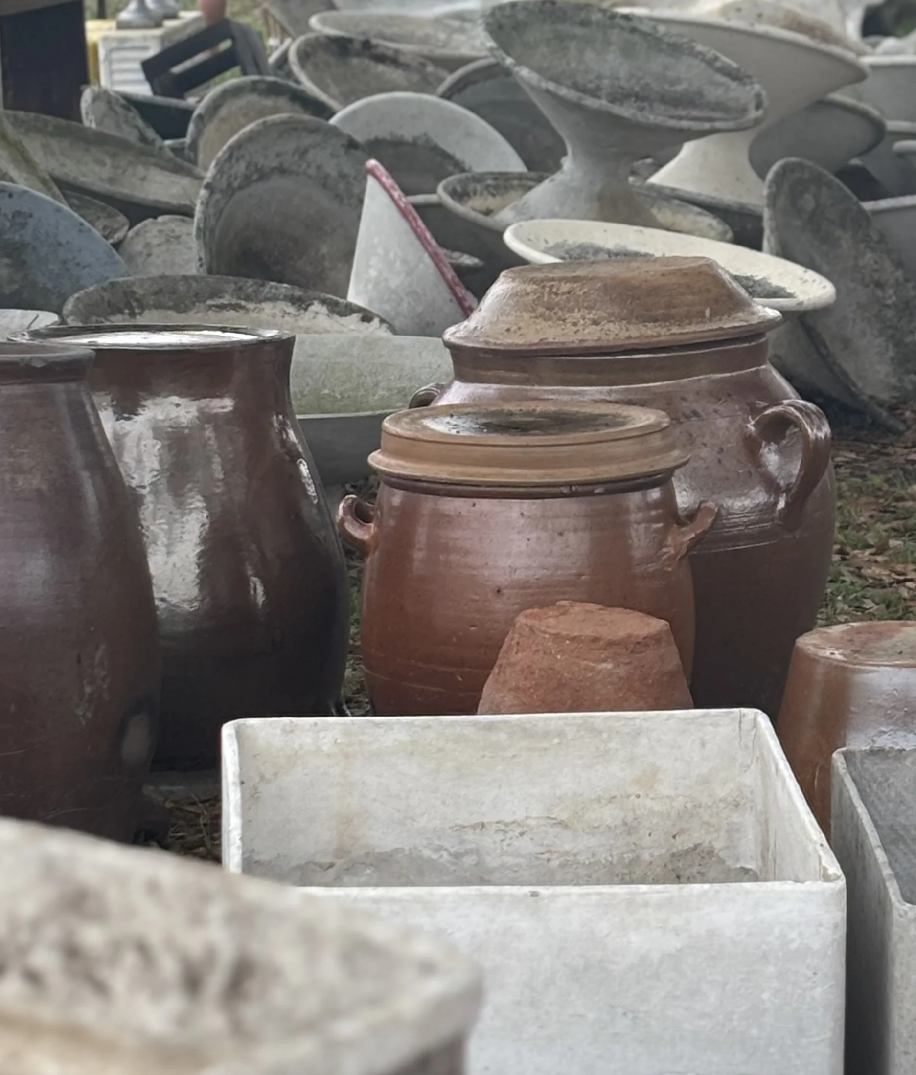 A collection of large ceramic pots and planters outdoors, some stacked and dusty, with a white rectangular container at the front, and a blurred background showing part of a person and other objects.