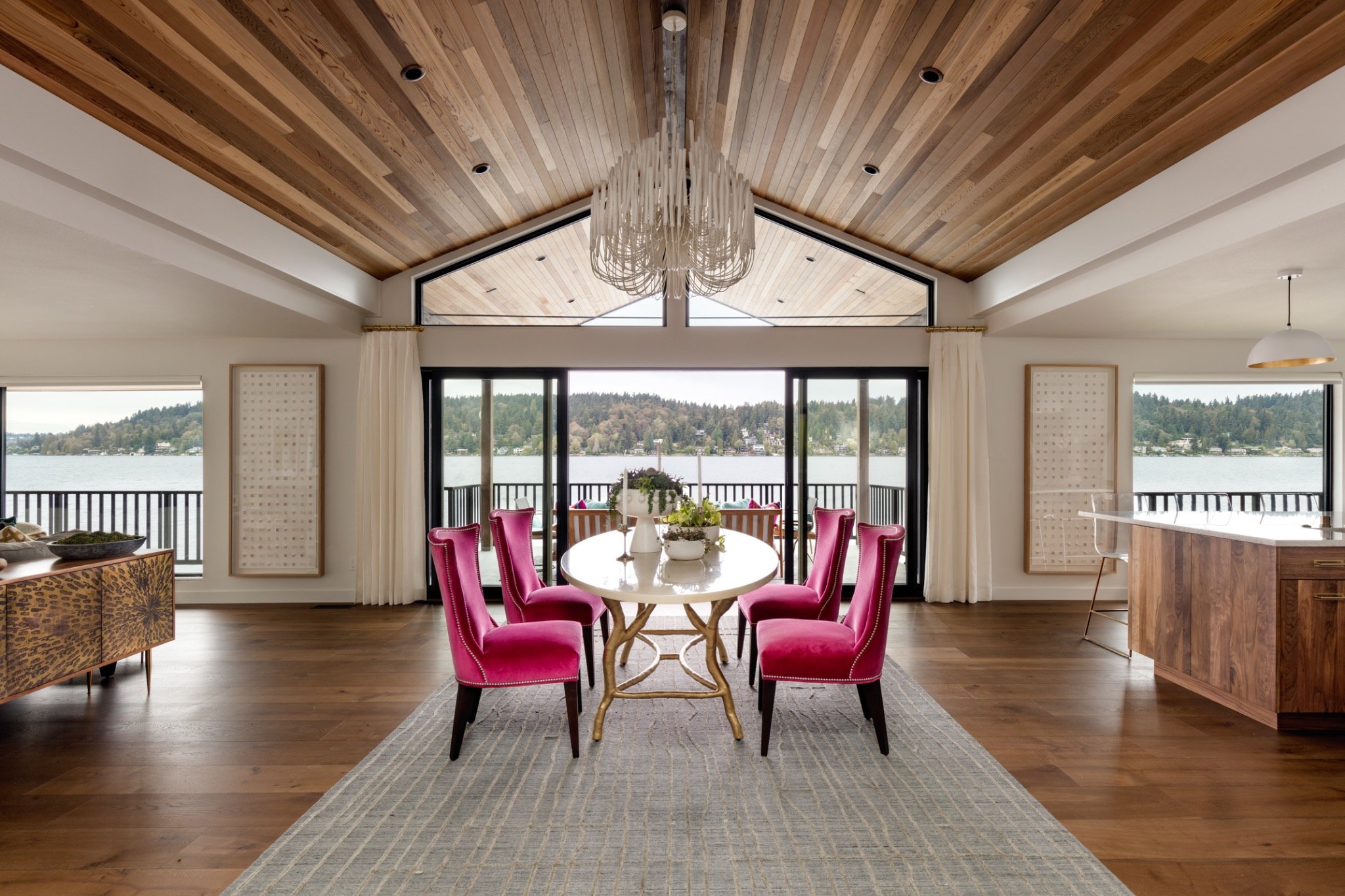 A dining room with a round table, six pink chairs, a chandelier, and large windows showing a lakeside view with trees and houses.