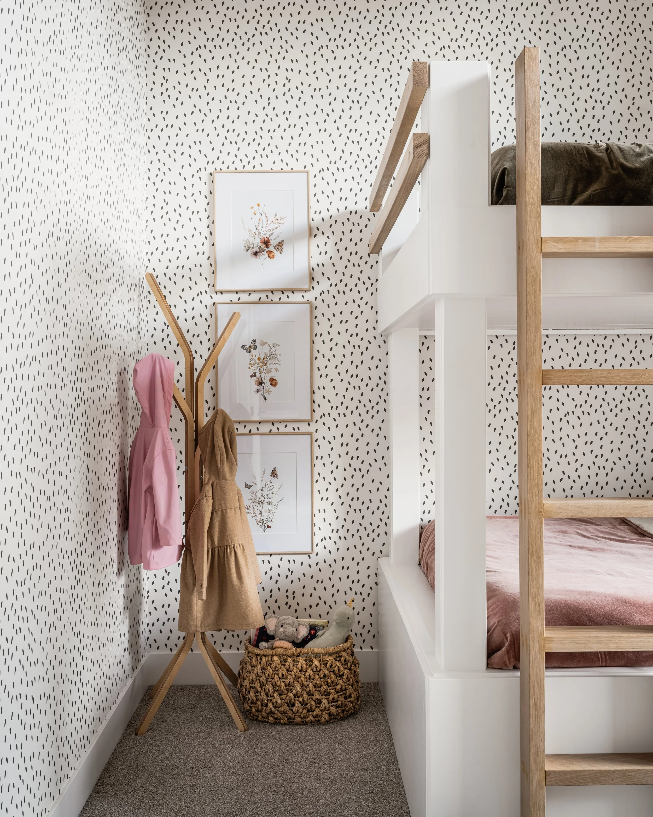 A small corner of a bedroom with a patterned black and white wallpaper, a framed botanical print, a wooden coat stand with pink and brown children's coats, a woven basket with stuffed animals, and a staircase with wooden railing leading to a lofted bed with dark bedding.