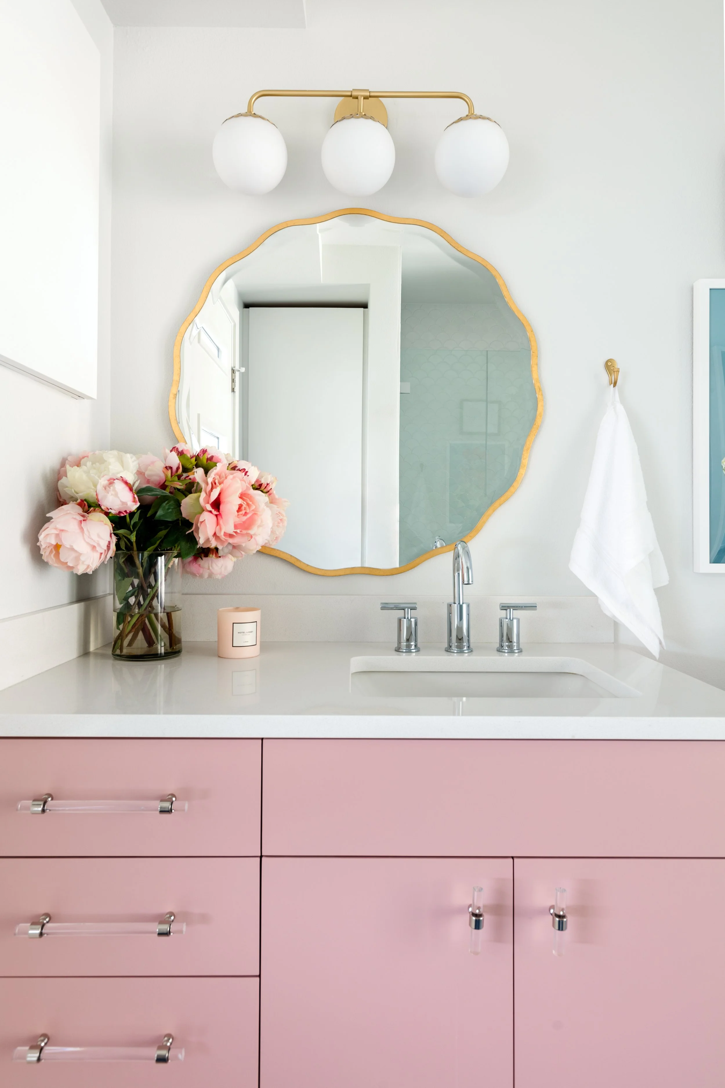 Bathroom vanity with pink cabinets, a white countertop, and a large mirror with a scalloped wooden frame. A bouquet of pink and white peonies, a pink candle, and a white towel hanging on a gold hook are on the counter. A three-light white and gold fixture is above the mirror.