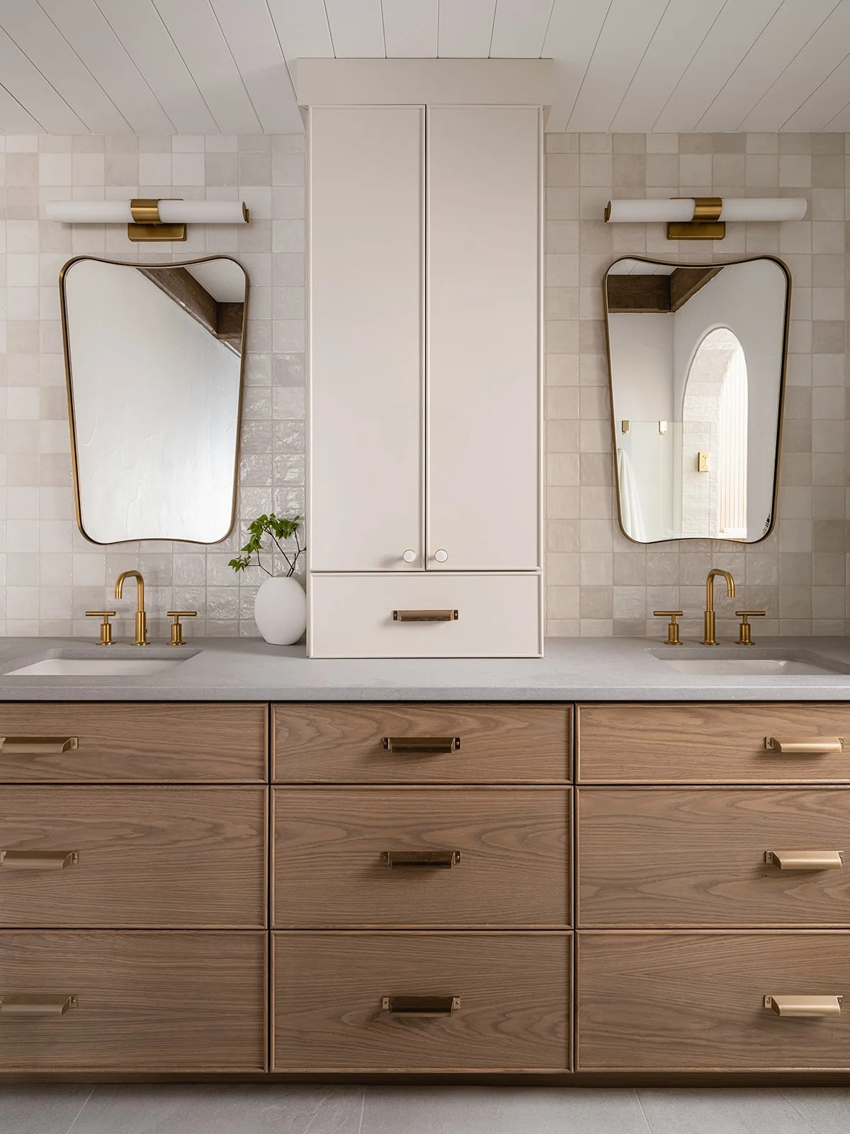 Modern bathroom vanity with two sinks, gold fixtures, and two mirrors, with a white cabinet in the middle and a potted plant on the left.