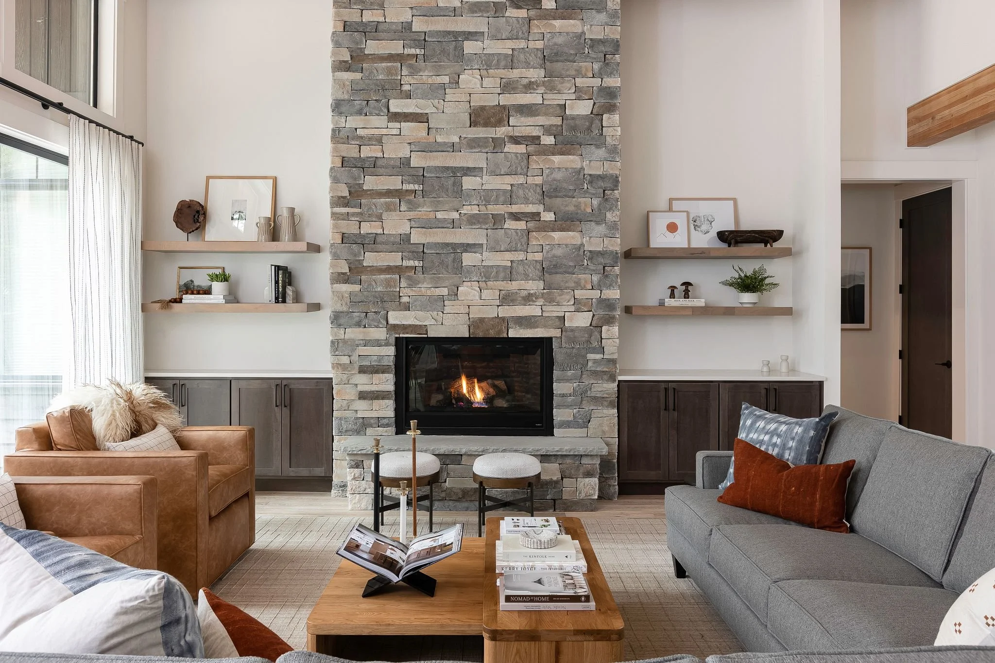 Living room with a stone fireplace, gray sofa with orange pillows, brown leather armchair with a cream pillow and throw, wooden coffee table with books and magazines, and floating shelves with decorative items and framed artwork. Large window with white curtains.