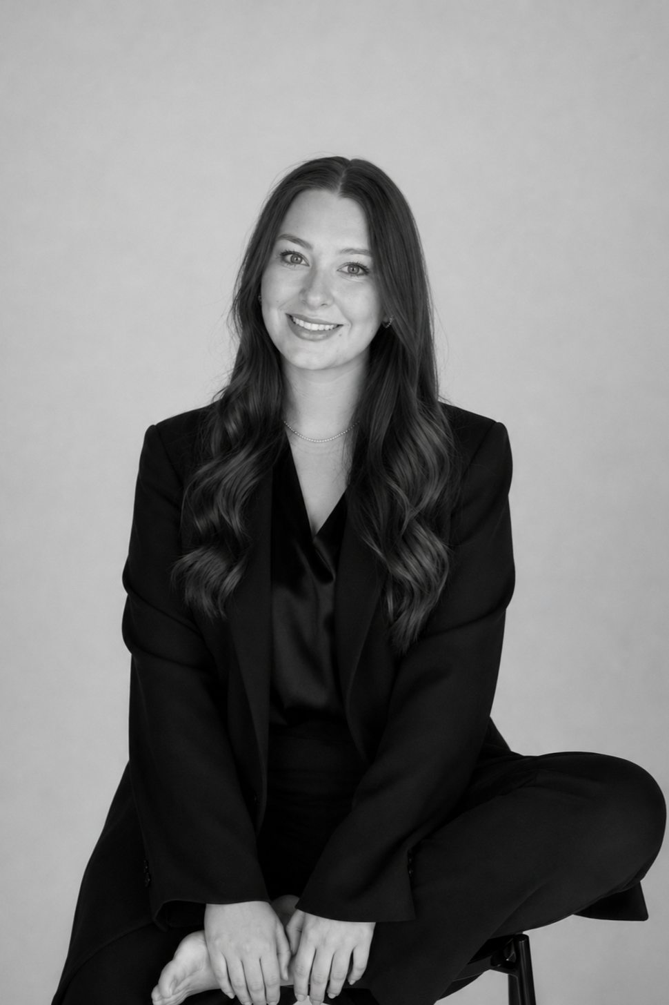 Black and white portrait of a young woman with long wavy hair, sitting cross-legged in a black blazer and top, smiling at the camera against a plain background.