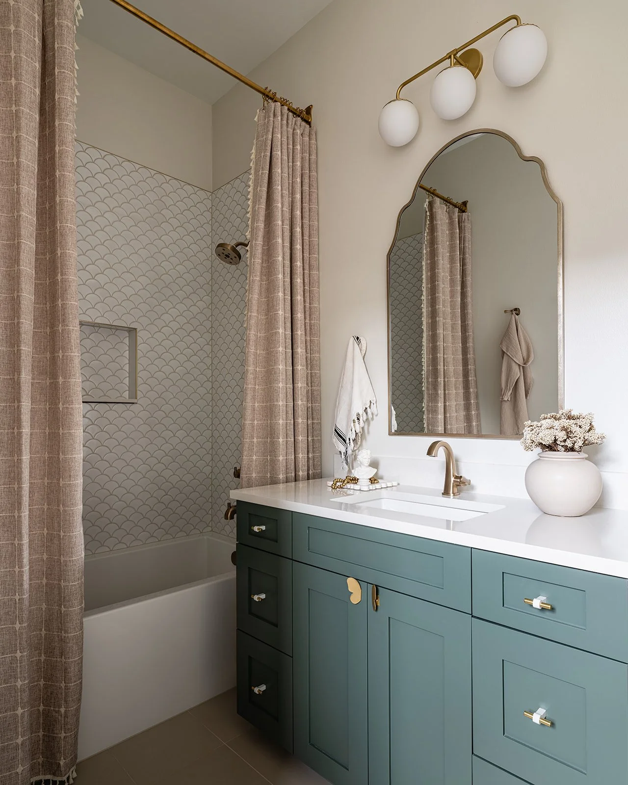 Modern bathroom with a teal vanity featuring gold hardware, a white countertop, a gold faucet, a large mirror with a gold frame, and a shower area with beige patterned curtains and tiles.