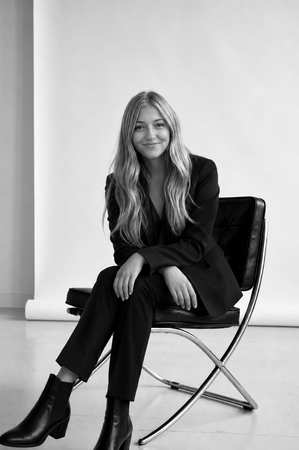 Black and white photo of a young woman with long wavy hair, sitting on a modern black leather chair, smiling at the camera, wearing a dark blazer, pants, and ankle boots, against a plain background.