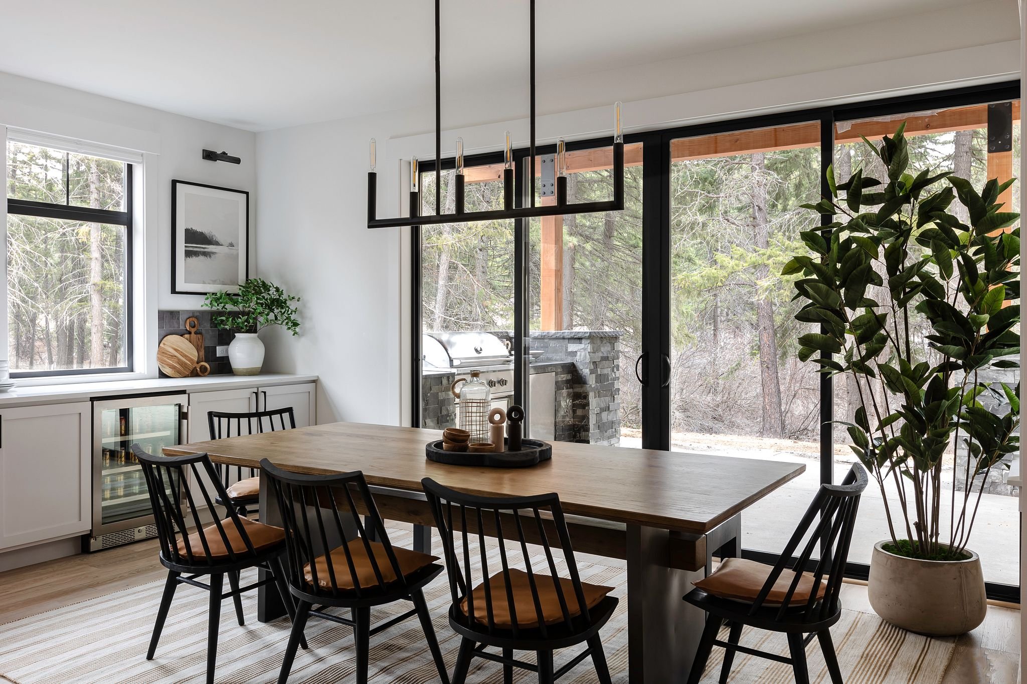 A modern dining room with a wooden table surrounded by black chairs with cushions. There is a large window and sliding glass door with a view of trees outside. The room has white walls, a hanging geometric light fixture, a framed black-and-white picture, a plant in a white pot, and a sideboard with jars and cutting boards.