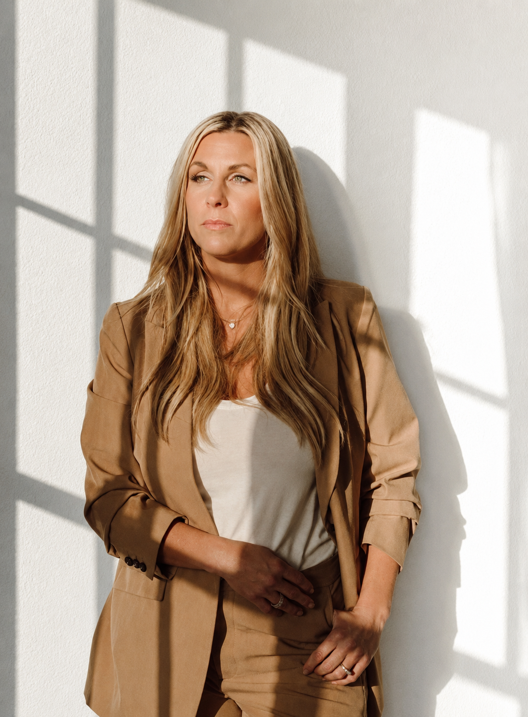 A woman with long blond hair wearing a beige suit and white shirt, standing against a white wall with sunlight creating shadows.