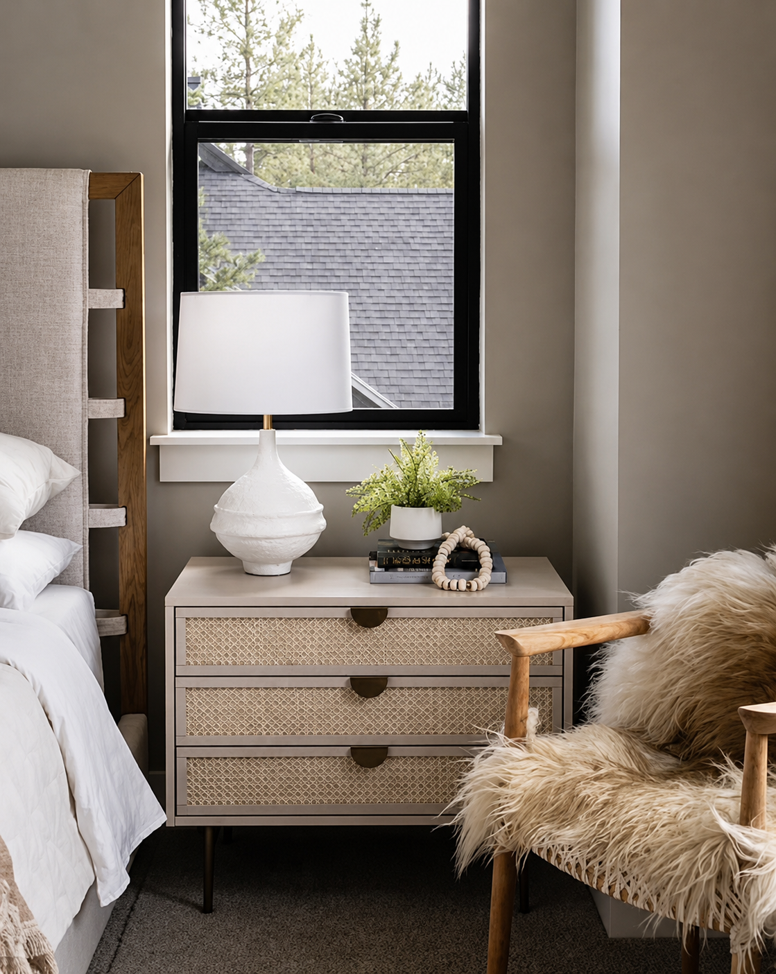 A cozy bedroom corner with a nightstand, white lamp, green plant, and decorative items, next to a window with a view of a house roof and trees outside.