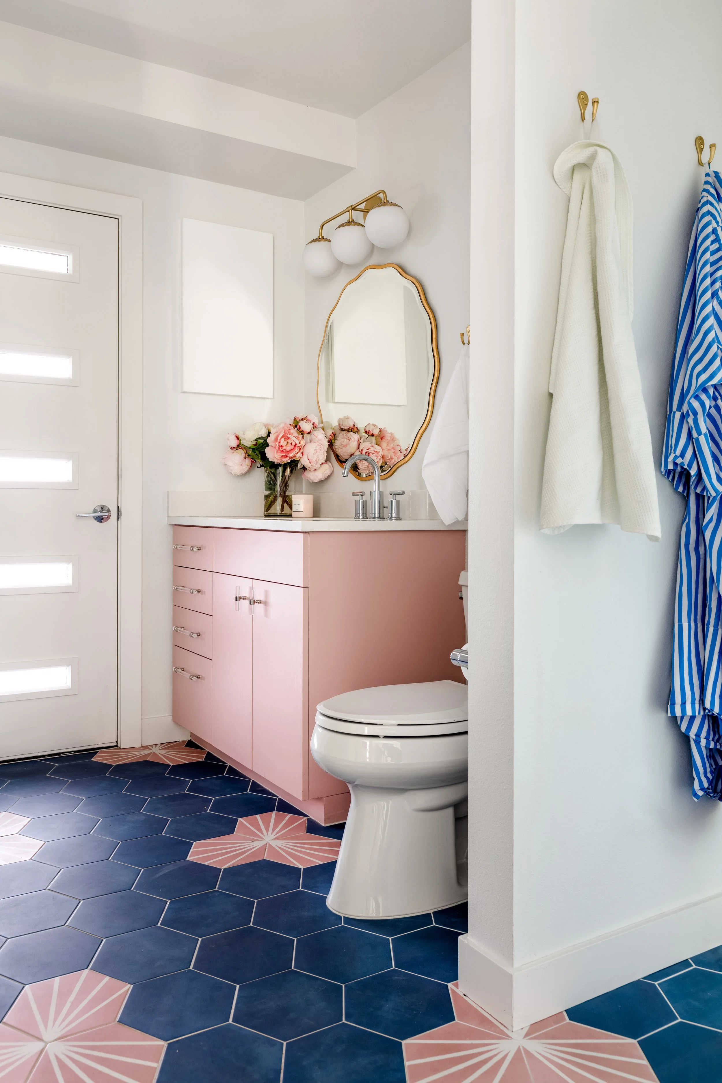 Bathroom with pink vanity, vase of pink flowers, mirror, white wall, blue and pink patterned floor tiles, white exterior door, hanging towels, and wall-mounted lighting fixture.