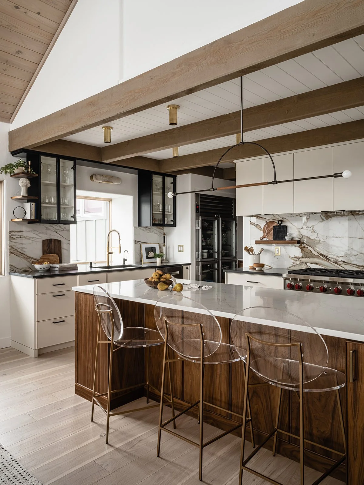 Modern kitchen with white marble countertops, wooden accents, and transparent barstools. Features include glass-front cabinets, a dual oven, and a large island with a fruit bowl.