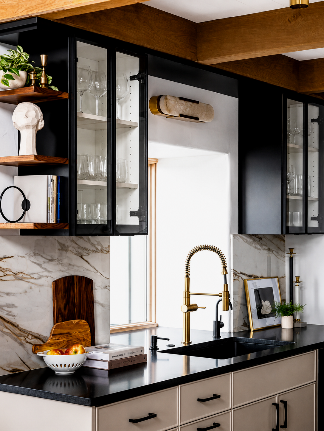 Modern kitchen with black and white cabinets, gold fixtures, marble backsplash, and open shelving with glassware, plants, and decorative items.