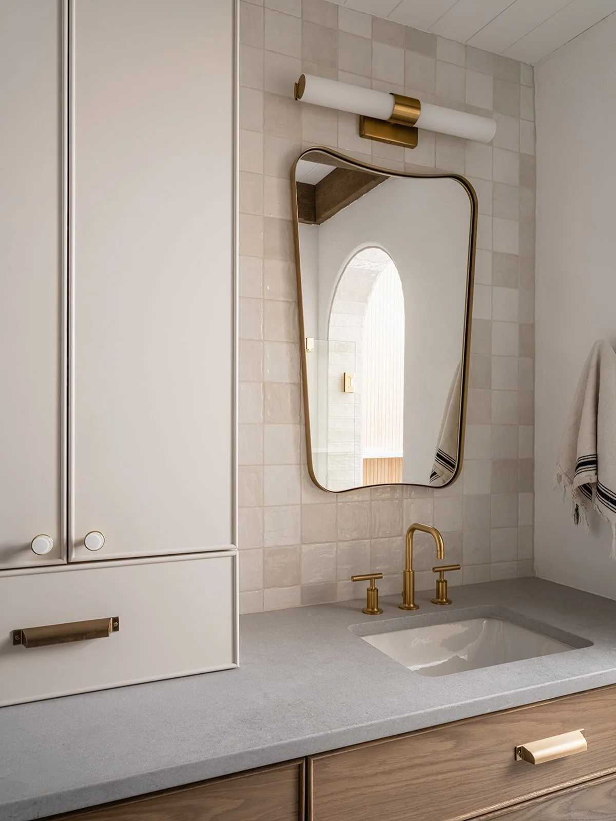 Bathroom vanity with a gold faucet, an oval mirror, and a white cabinet with gold handles, beige tiled wall, white countertop, and a gold wall-light fixture.