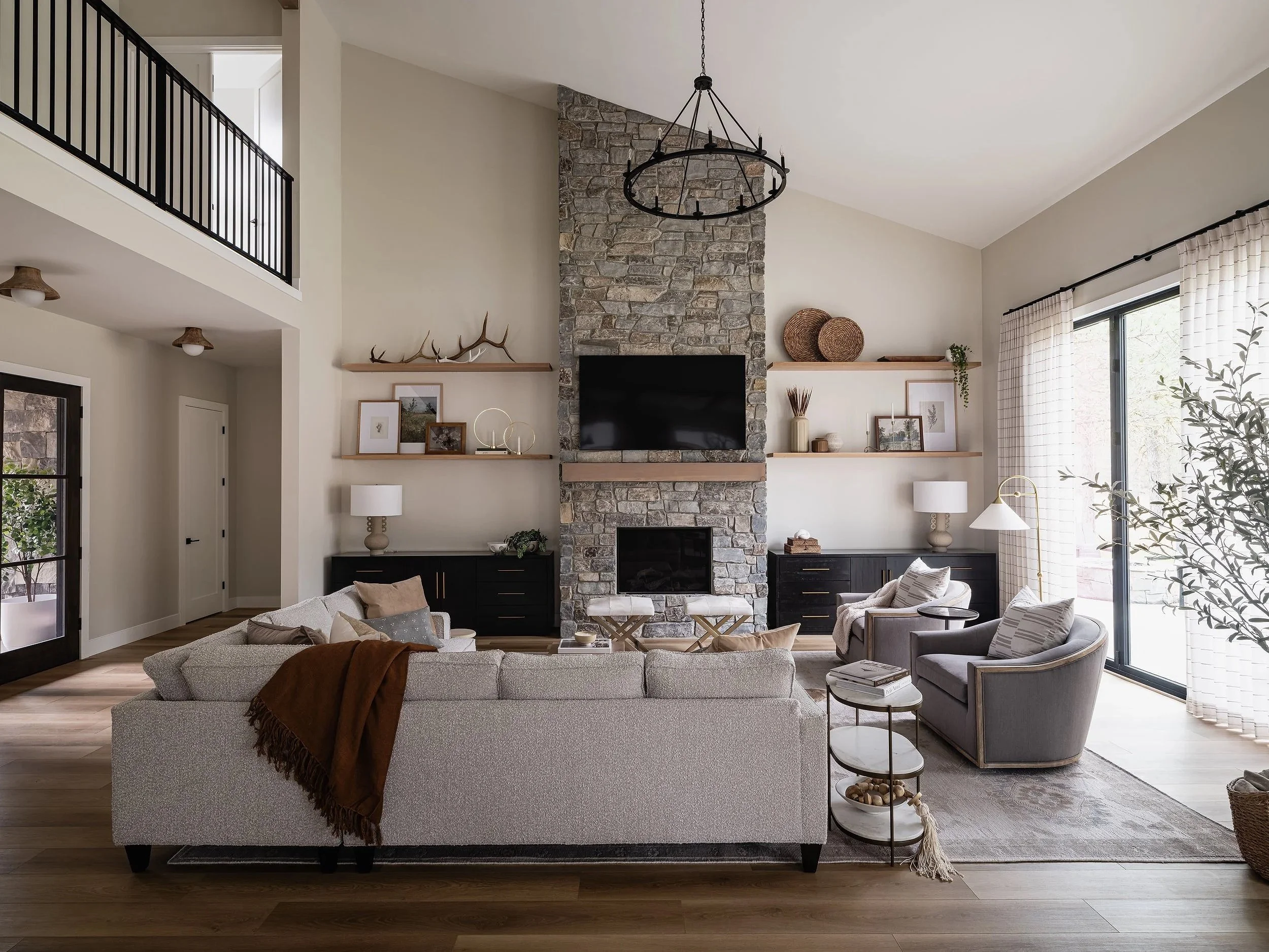 Living room with a stone fireplace, TV, beige sofa with pillows, modern side tables, and large windows with curtains.