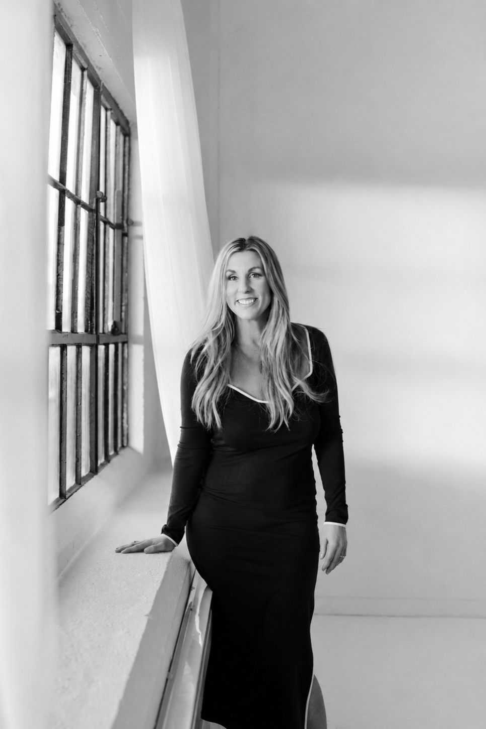 A woman in a long black dress standing by a window with metal bars, smiling and looking at the camera, in a bright room with light-colored walls.