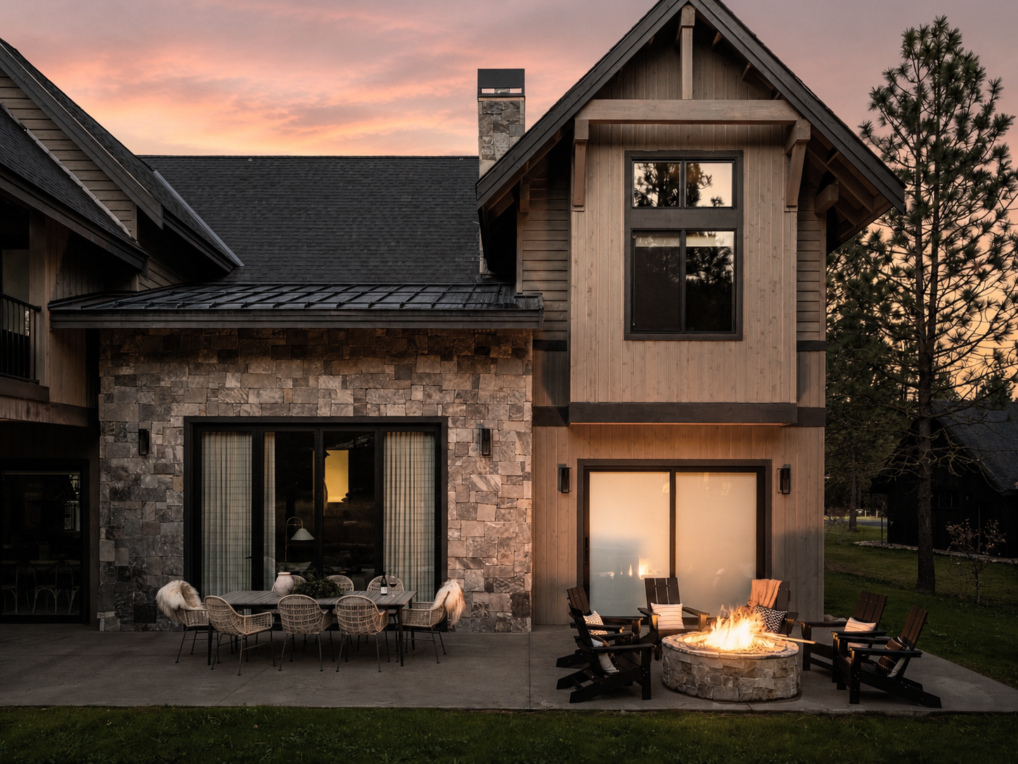 Exterior view of a modern house at sunset with a stone and wood facade. The backyard features a dining table with six chairs, and a fire pit surrounded by five Adirondack chairs, with the house's large windows reflecting the sunset sky.