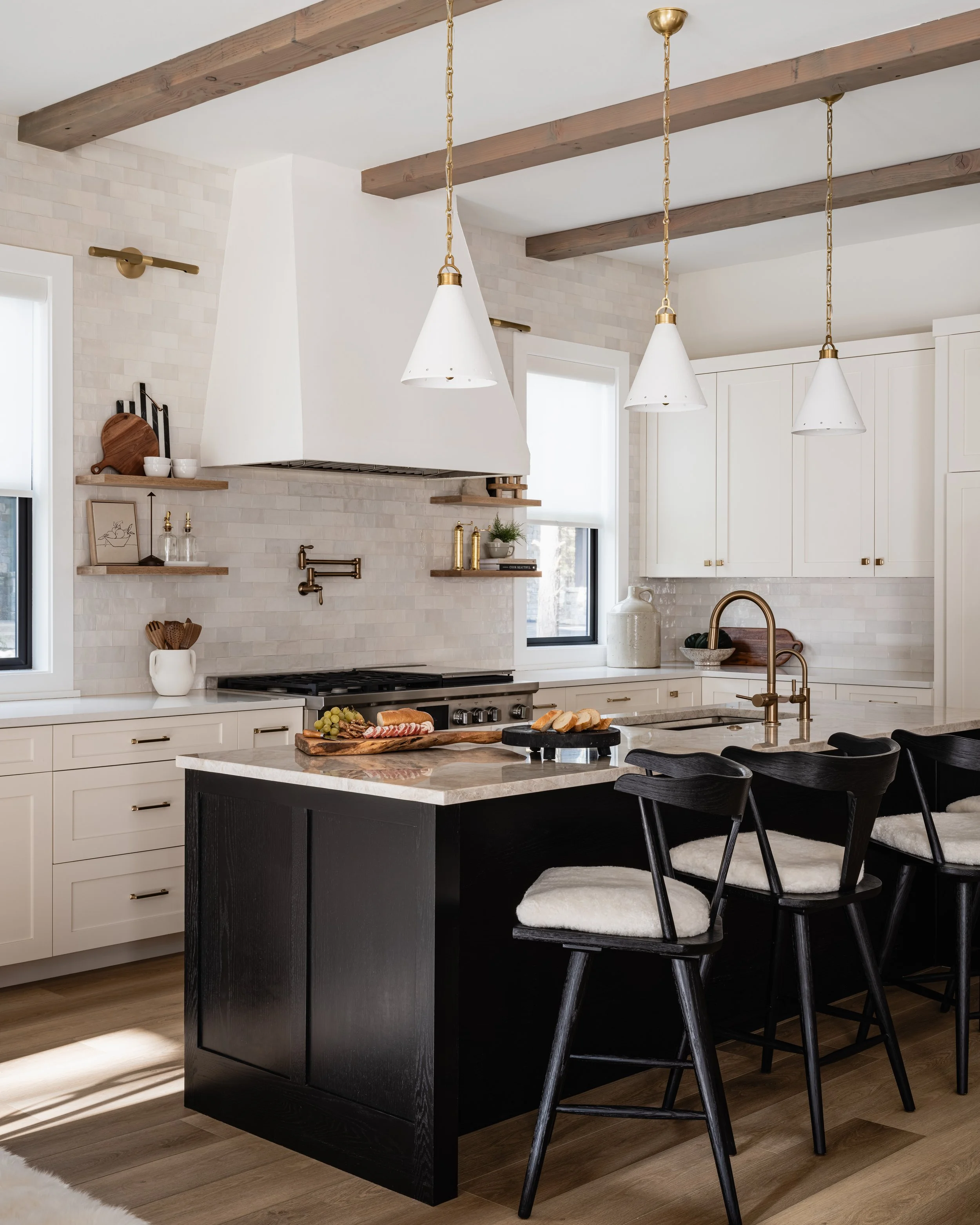 Modern kitchen with a black island, white cabinets, and hanging white cone-shaped pendant lights with gold accents. The kitchen has wooden ceiling beams and light-colored walls.