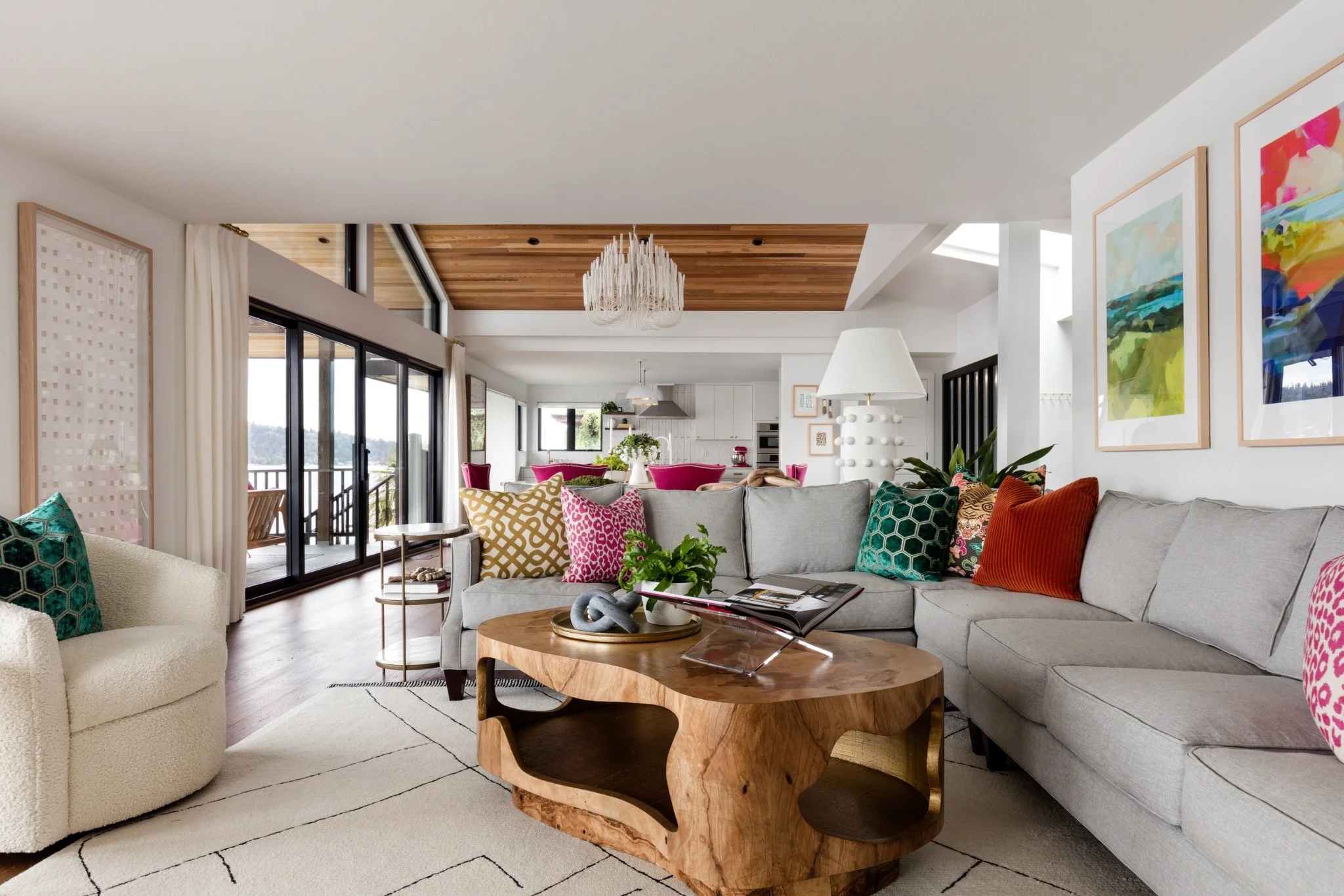 Bright living room with a gray sectional sofa adorned with colorful patterned pillows, a wooden coffee table, large windows with curtains, and artwork on the walls. The space extends into a kitchen and dining area with a wooden ceiling and pendant lighting.