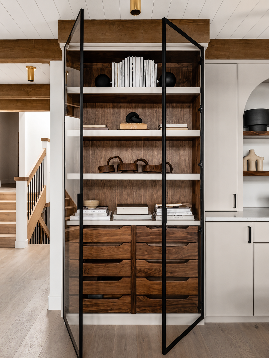 Wood and white shelving cabinet with books, decorative objects, and drawers, enclosed in a glass door, in a modern home interior.