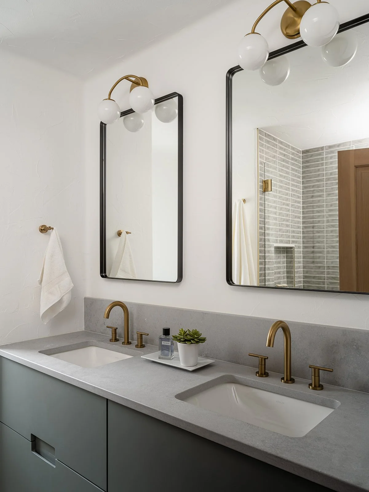 Modern bathroom with a double sink vanity, gold fixtures, two rectangular mirrors with black frames, white walls, a soap dispenser, a small green potted succulent, and two light fixtures with white globes.