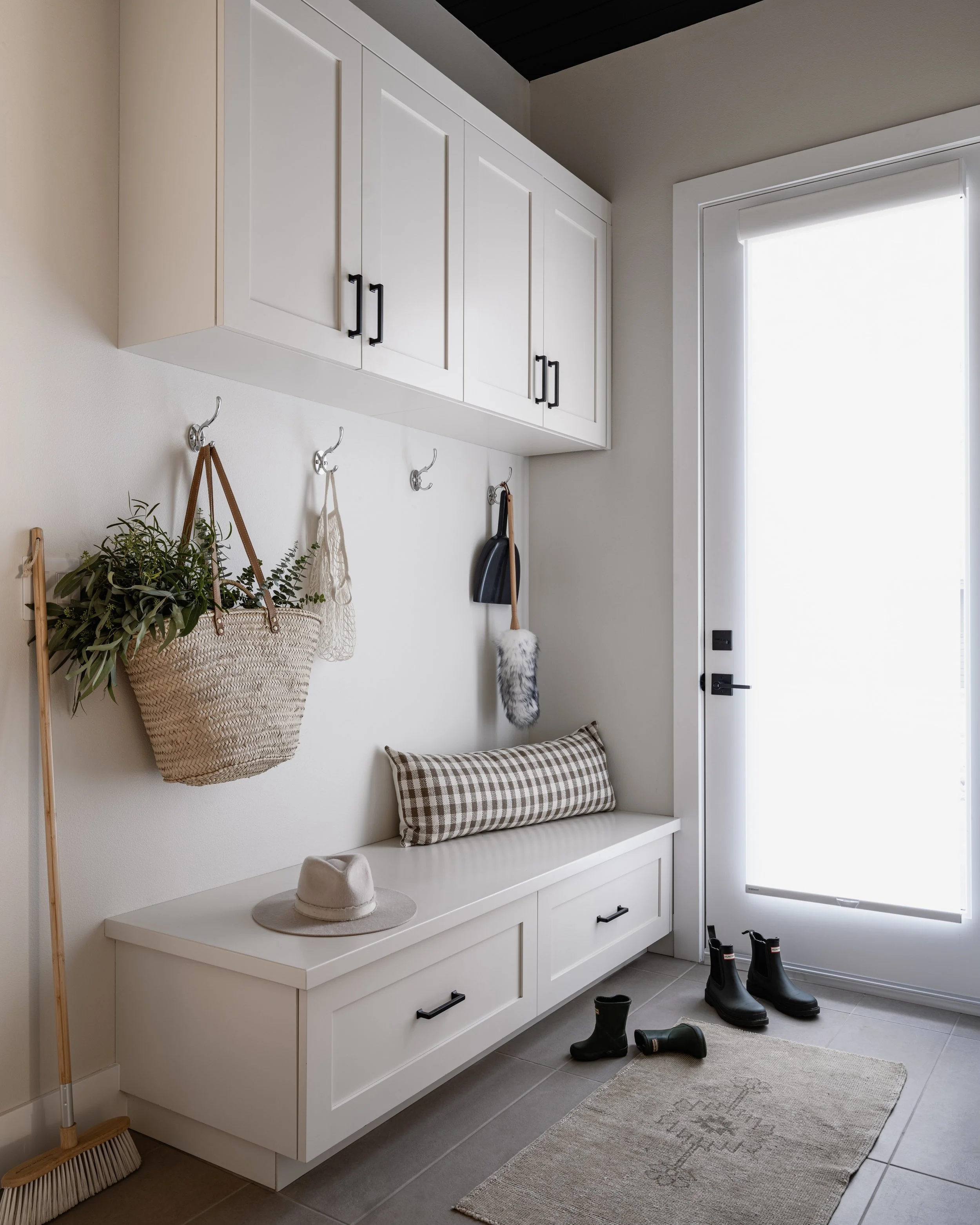 Entryway with white built-in storage bench, overhead cabinets, a row of coat hooks with hanging plants and accessories, black rain boots, a beige hat on the bench, a patterned pillow, and a light gray rug, next to a white door with a window.