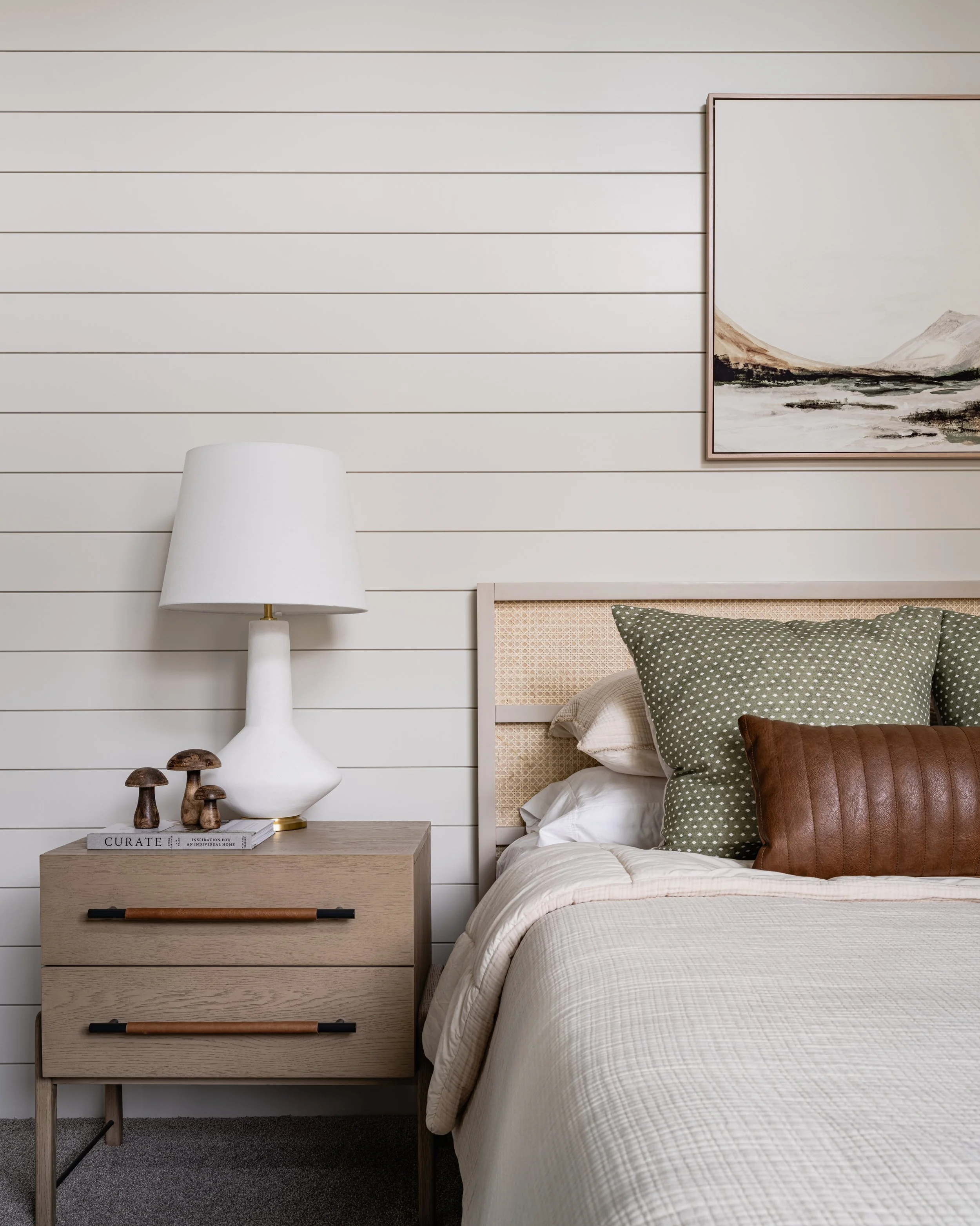 A bedroom with white horizontal shiplap walls, a bed with beige checkered bedding, multiple pillows including dark green and brown ones, a wooden headboard, a wooden nightstand with a large white lamp, mushrooms, and a book titled 'Curate'. A framed landscape painting hangs on the wall.