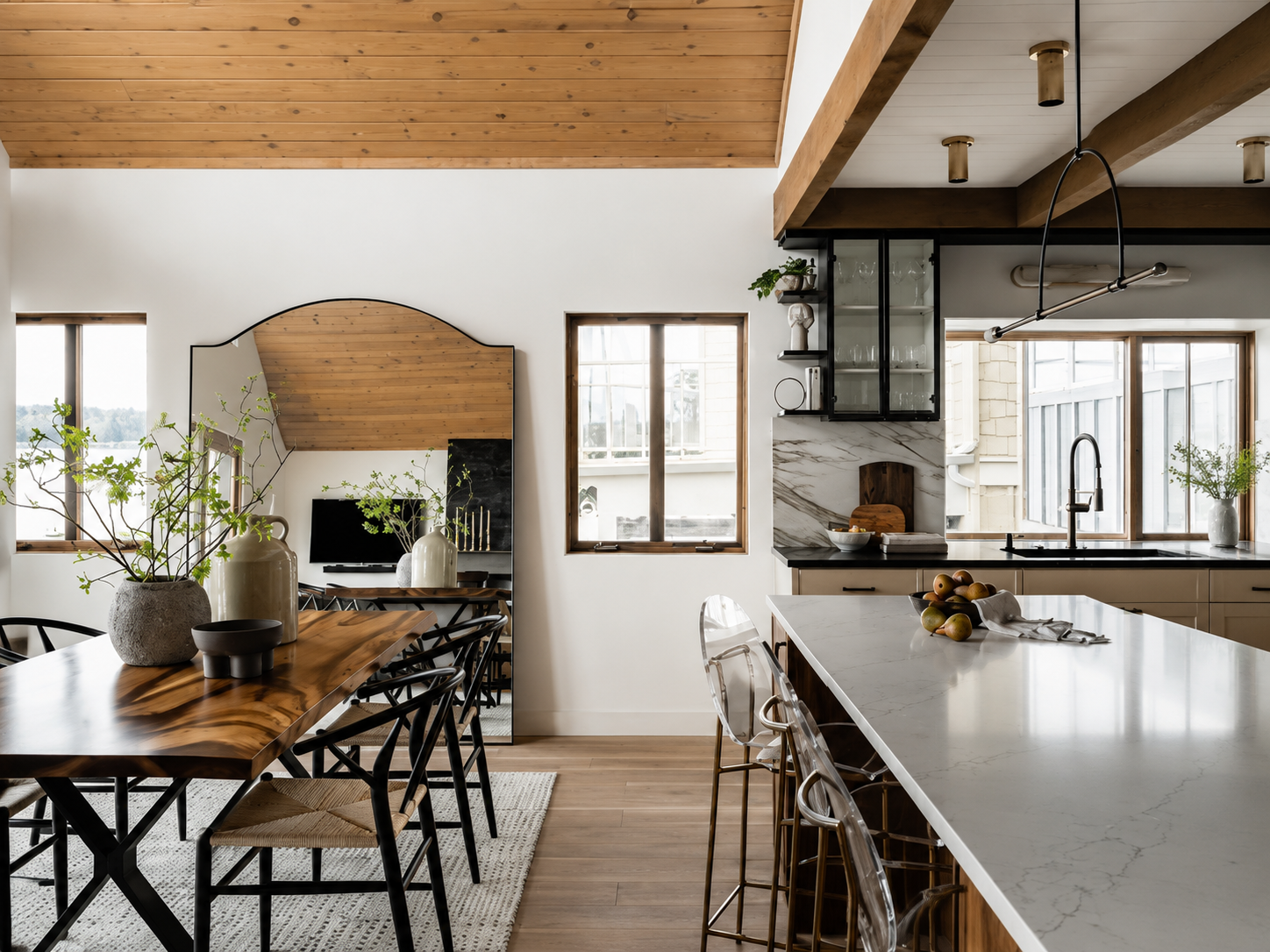 Modern kitchen and dining area with wooden ceiling and white walls, featuring a marble-topped island and a wooden dining table with chairs. Decor includes potted green plants and vases.