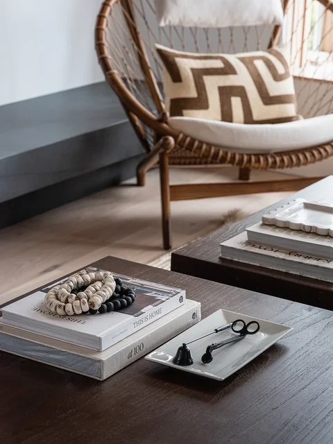 Living room corner with a round wicker chair, beige and brown patterned pillow, dark wood coffee table with stacked books and decorative objects, and a light-colored wooden floor.