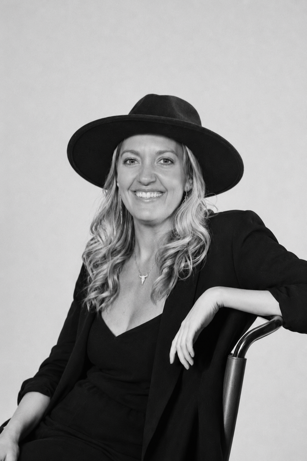 A woman with long curly hair wearing a wide-brimmed hat, black blazer, and black top, smiling while sitting on a chair, in black and white.