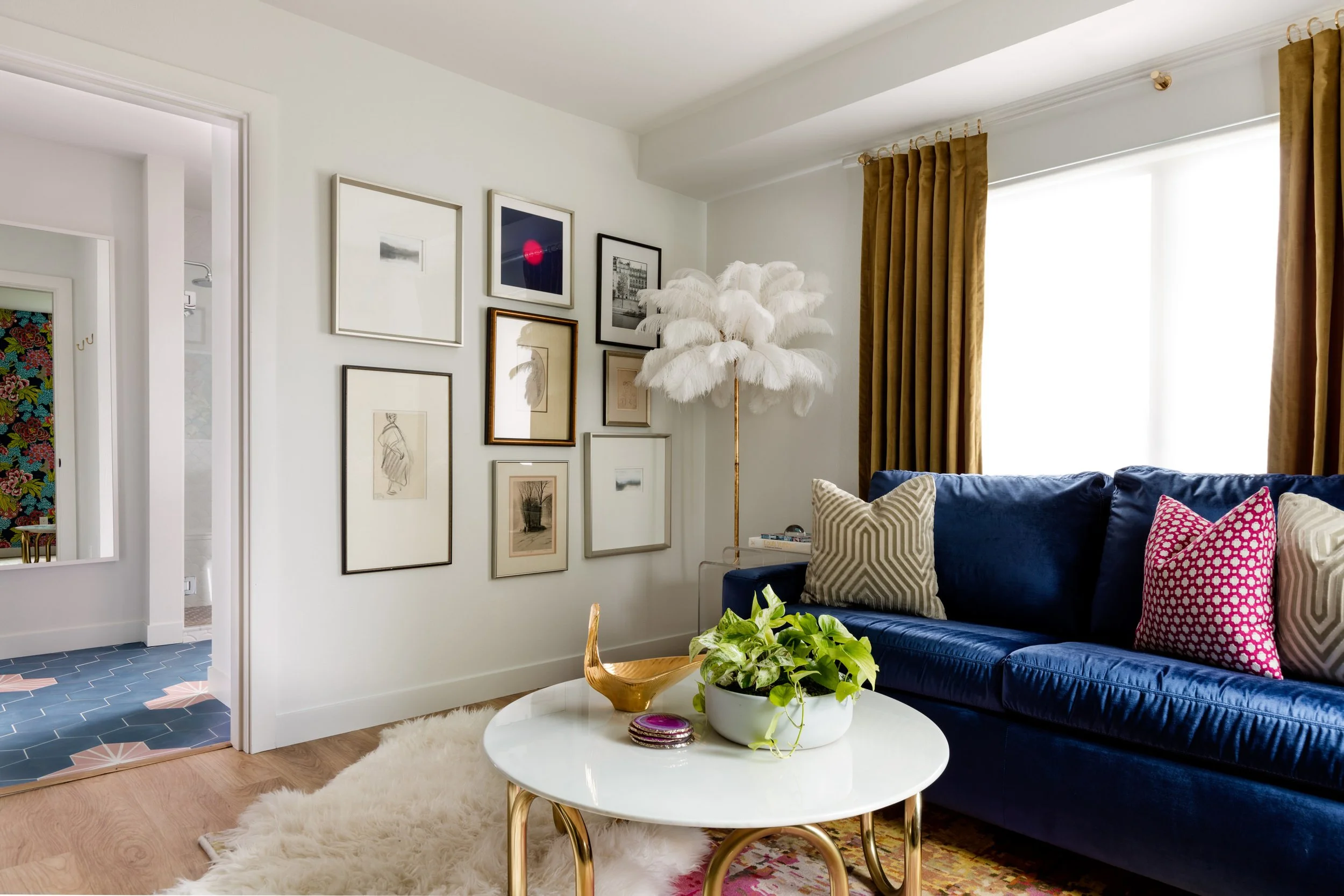A living room with a navy blue velvet sofa decorated with patterned throw pillows, a white wall adorned with various framed art pieces, a white fluffy feather lamp, gold curtain, a white coffee table with a potted plant, and a window allowing natural light to fill the room.