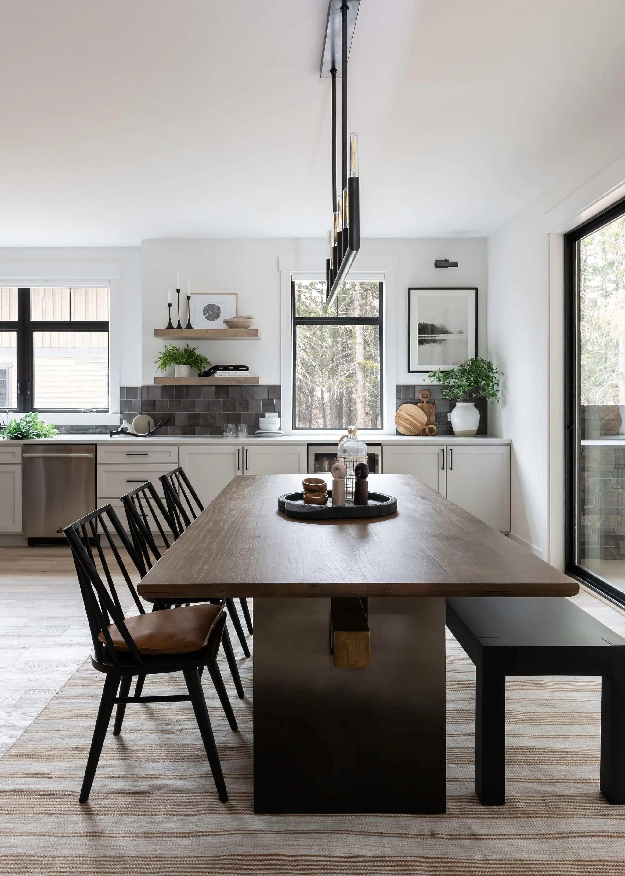 Modern kitchen with white cabinets, black accents, a wooden dining table, black chairs, and large windows showing trees outside.