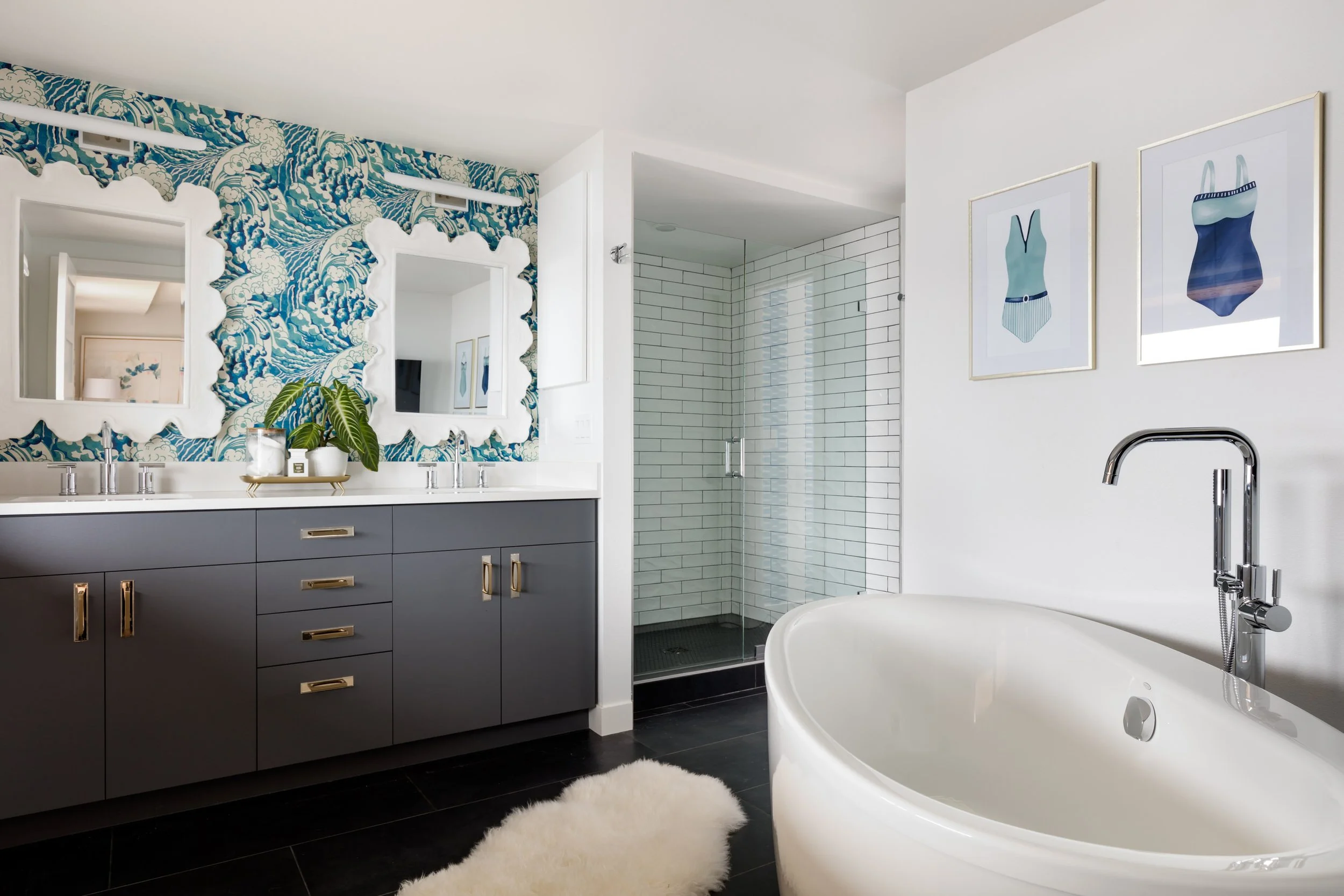 Modern bathroom with a blue vanity, frameless mirrors, a walk-in shower with white subway tiles, a white bathtub, and framed artwork of women wearing swimsuits on the wall.