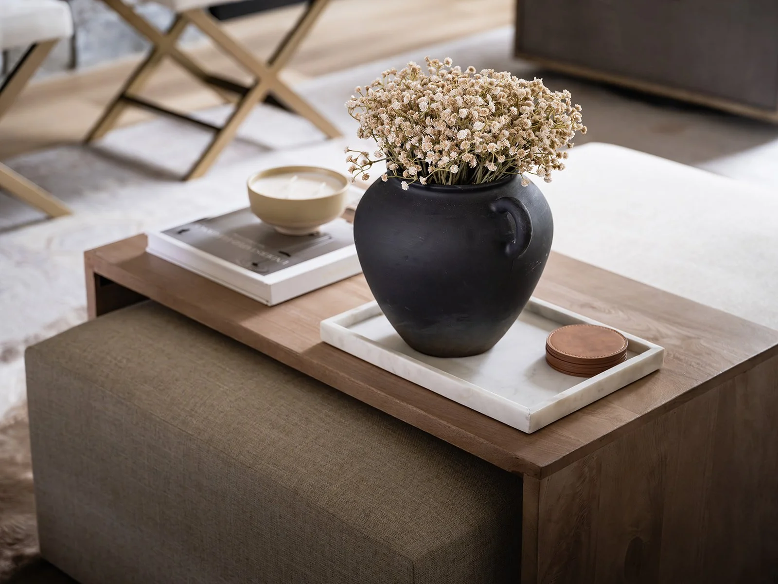 A black vase filled with white dried flowers on a wooden coffee table, with a tray holding a white bowl, some coasters, and magazines or books in a modern living room.