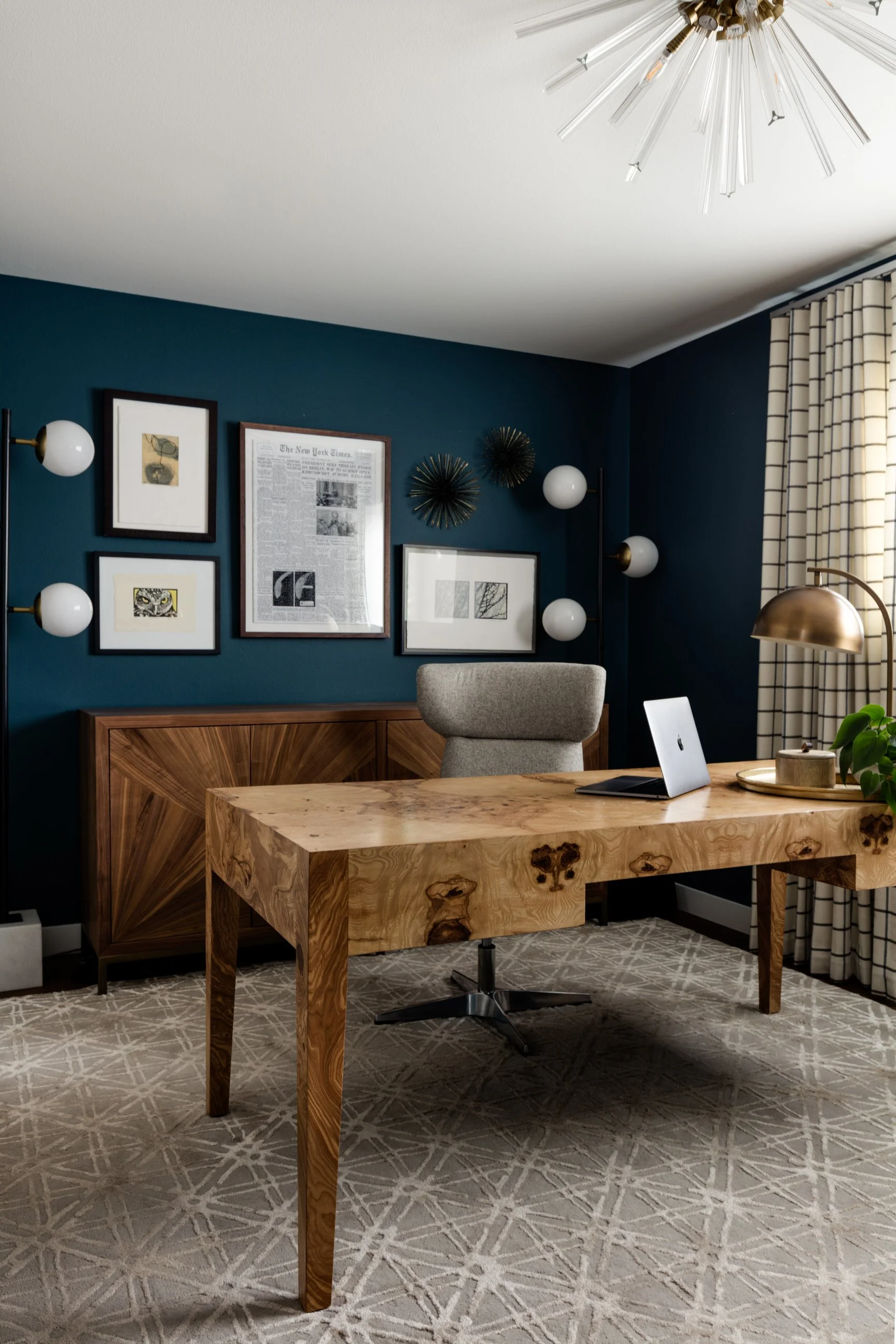 A modern home office with a wooden desk, a gray office chair, and a computer. The wall is dark teal with framed pictures and decorative wall art. A window with plaid curtains is on the right, and a gold desk lamp and a floor lamp with globe bulbs are present. The ceiling has a unique modern light fixture.