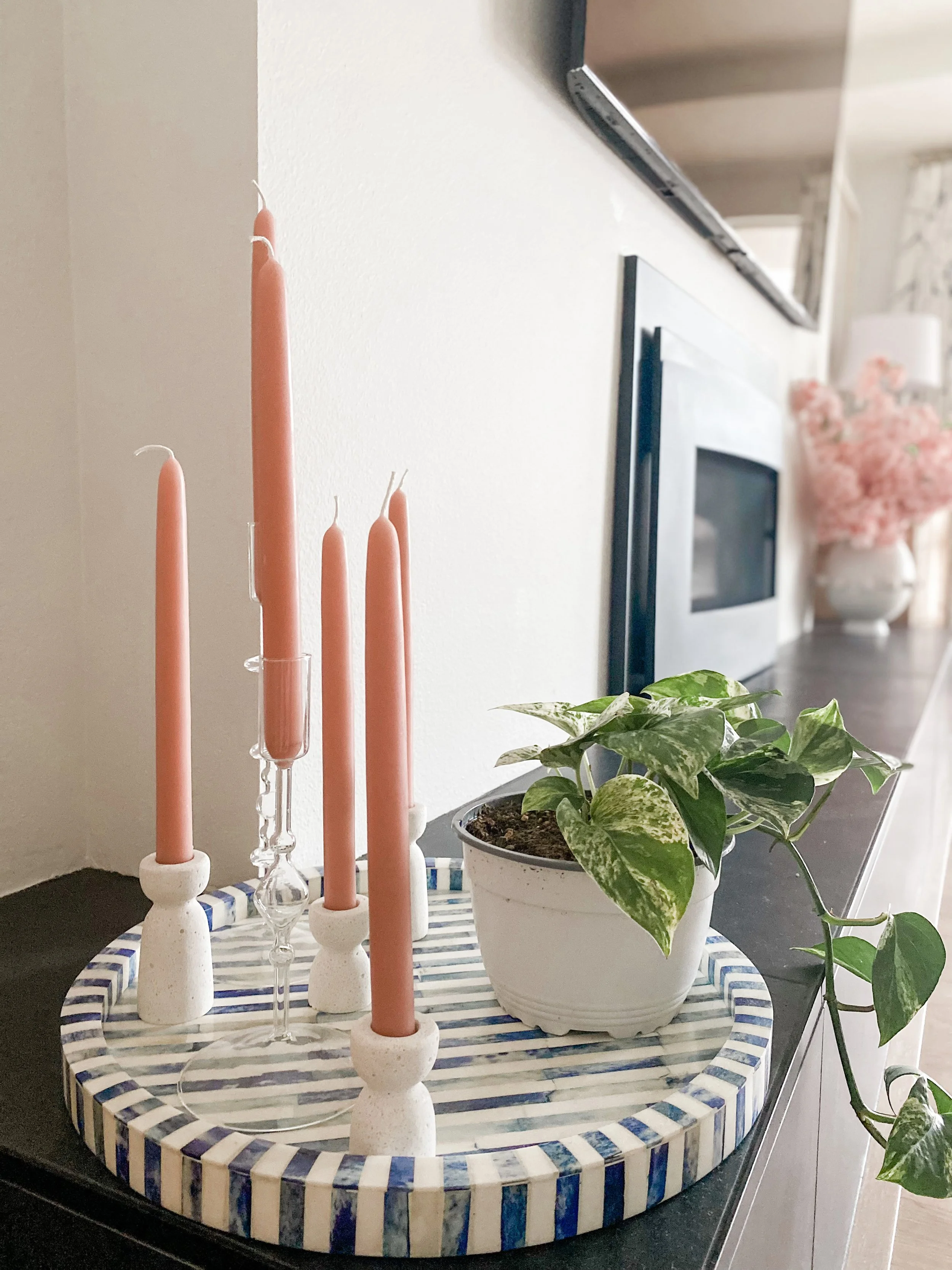 A decorative table display featuring five pink candles on ceramic holders, a potted green plant with variegated leaves, and a large vase with pink flowers in the background, set on a striped blue and white tray placed on a black surface.