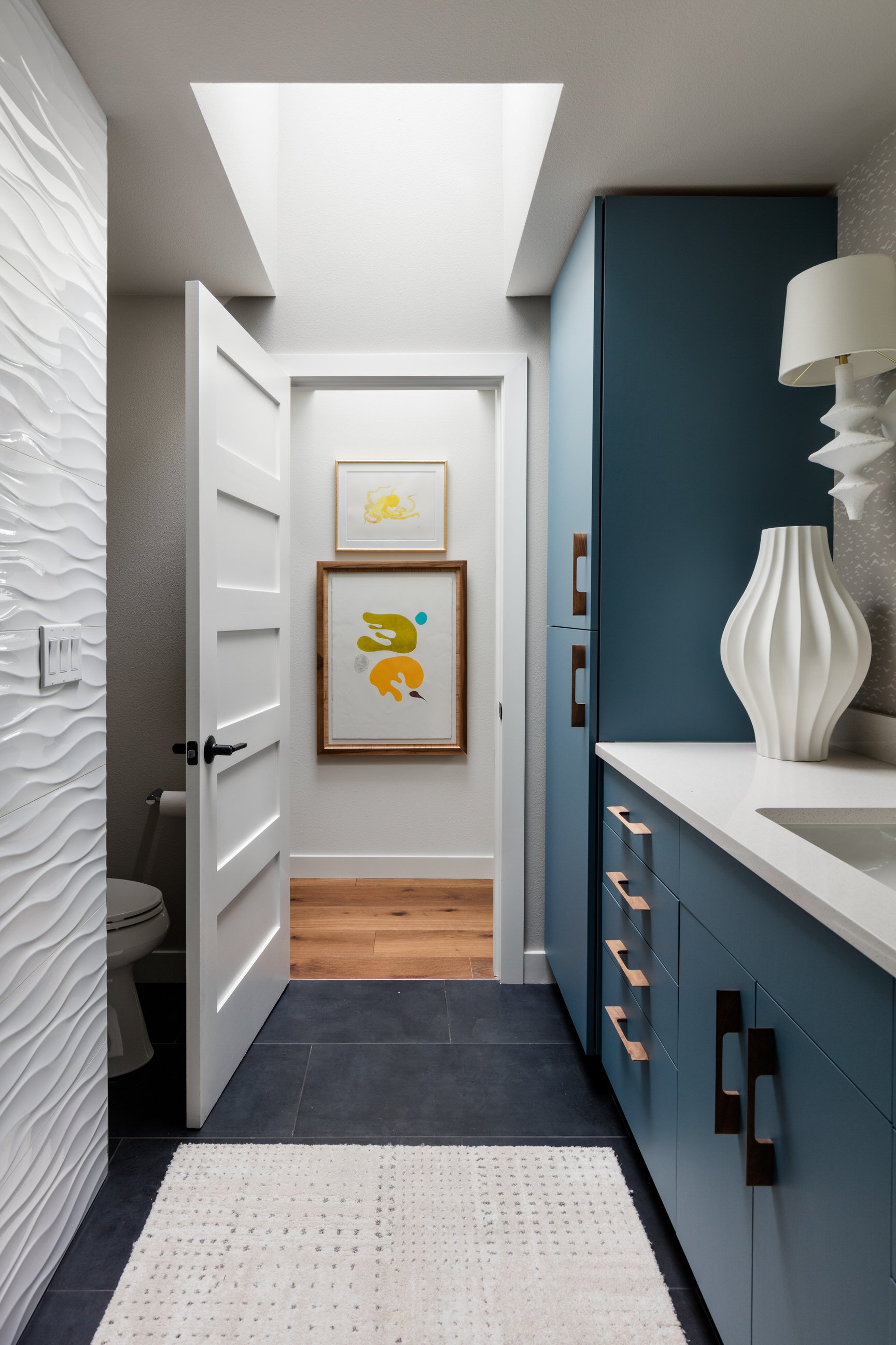 A modern bathroom with dark gray floor tiles, a white textured wall on the left, and a blue vanity with wooden handles on the right. There is a white countertop with a white sculptural table lamp. At the back, artwork featuring abstract shapes and a yellow lion is visible on the white wall beyond an open door. The ceiling has a skylight allowing natural light in, and there is a beige rug on the floor.