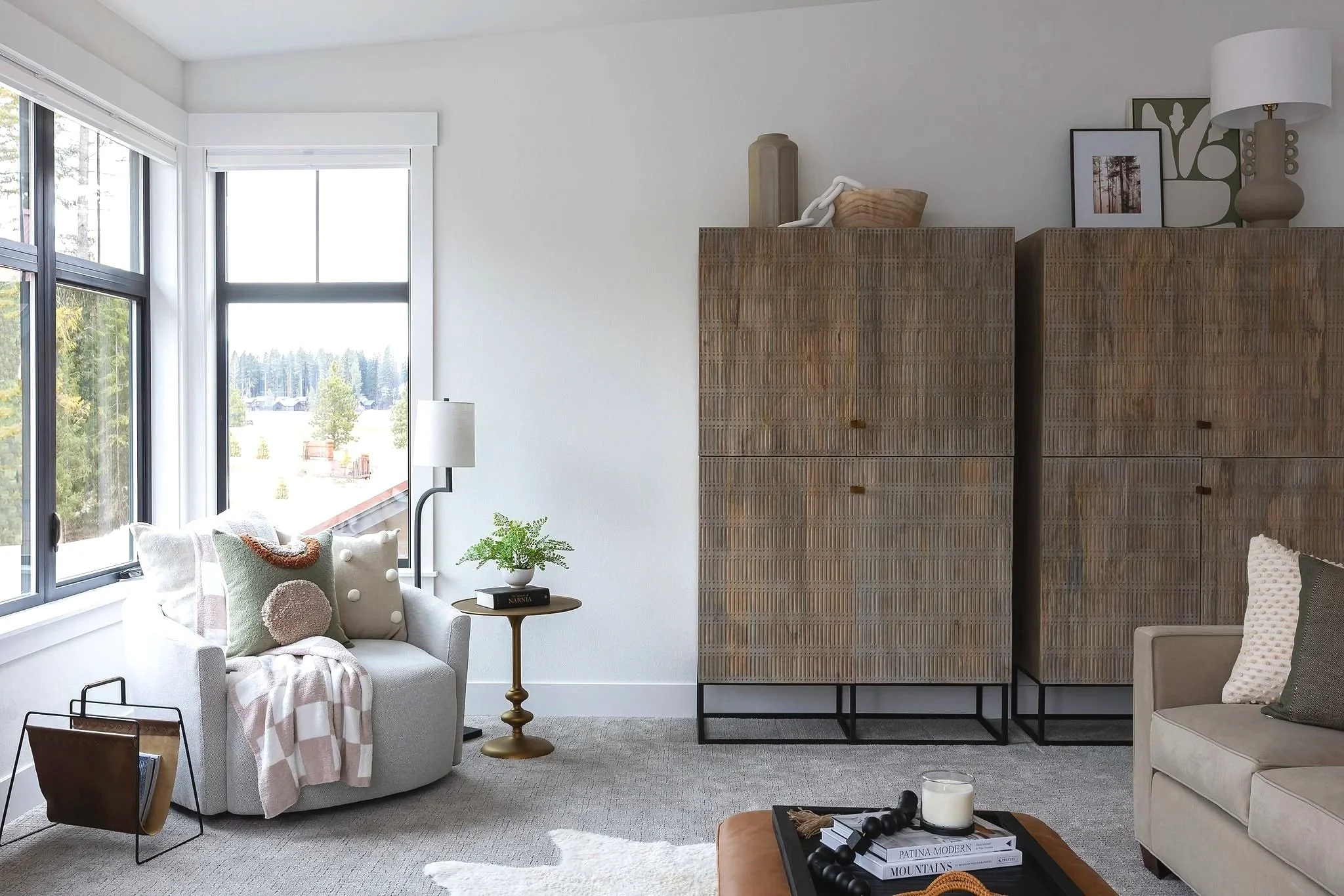 A modern living room with large windows, beige sofas with pillows, a small round side table with a plant and books, a tall wicker room divider, artwork, a table lamp, and a coffee table with decorative objects, all in a bright and airy space.