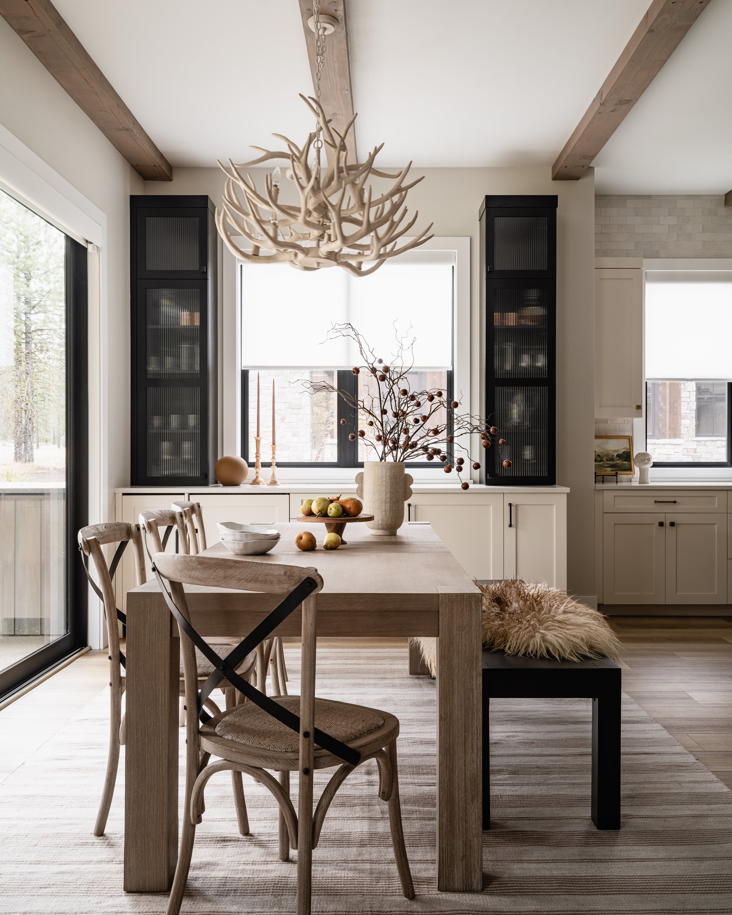 A dining room with a wooden table, chairs, a chandelier made of antlers, a table centerpiece with branches and decorative balls, and natural light coming through large windows.