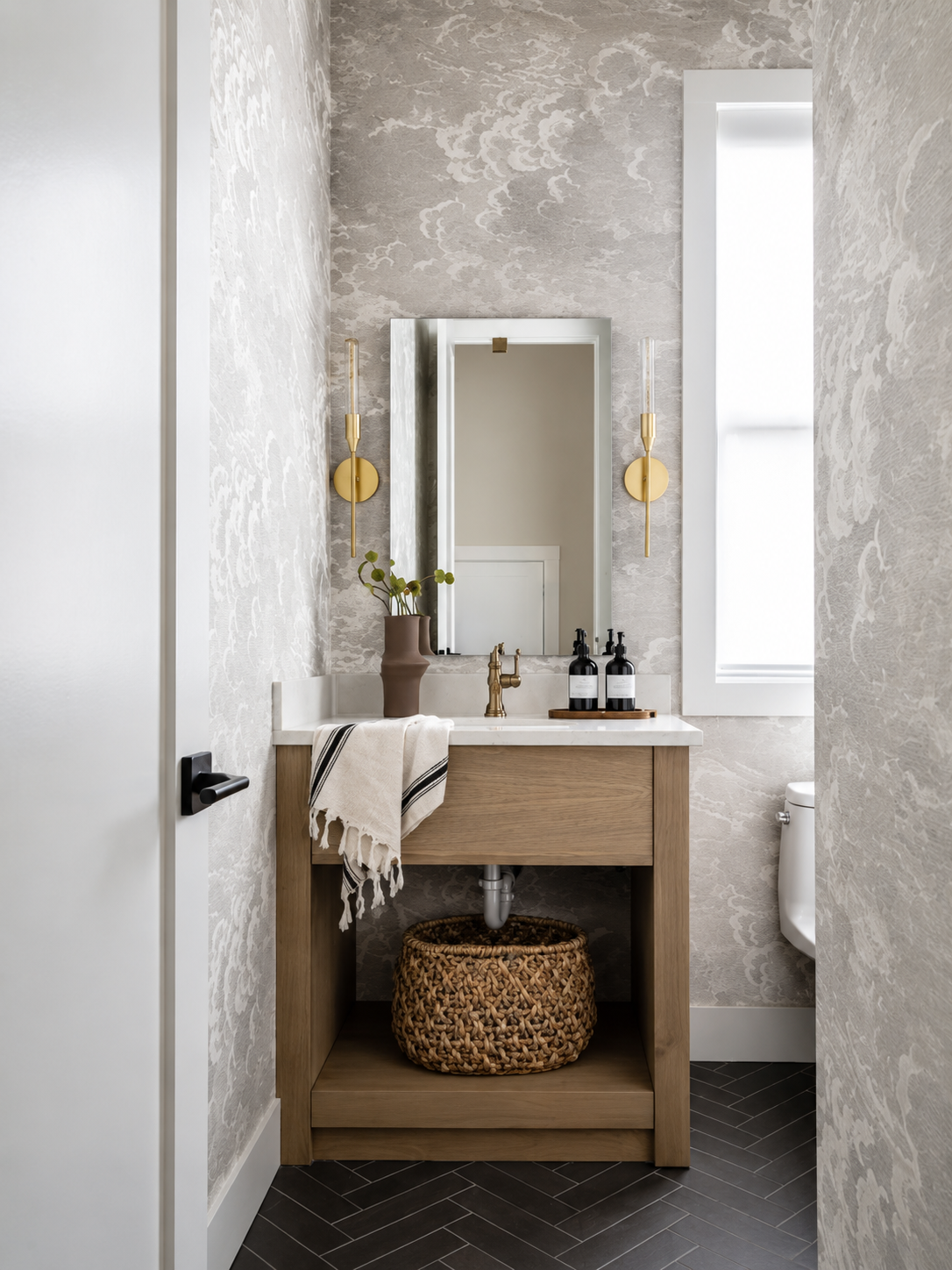 Small bathroom with a vanity, framed mirror, beige wallpaper, wall-mounted gold sconces, a window with white trim, and a woven basket underneath the sink.